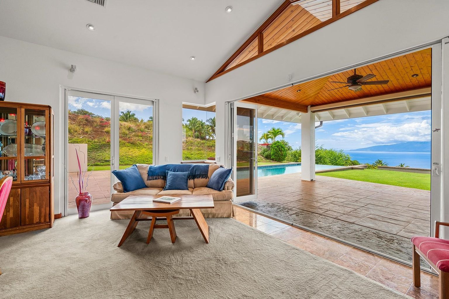 This is an interior shot of a living room with a beige sofa adorned with blue pillows, a wooden coffee table, and a display cabinet. The room opens to an outdoor patio with a pool and ocean view through large sliding glass doors, creating a seamless indoor-outdoor living experience. The vaulted ceiling with wooden beams adds architectural interest.