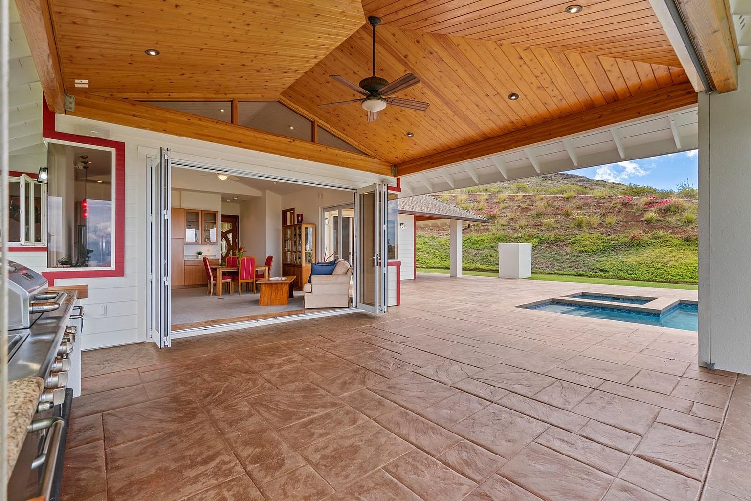 This image showcases a spacious outdoor patio area with a stamped concrete surface, seamlessly connecting the indoor and outdoor living spaces through large, folding glass doors. The patio features a wooden ceiling with a ceiling fan, and a built-in pool/spa is visible in the background, set against a lush green hillside. The design promotes an open and airy atmosphere, perfect for entertaining or relaxing.