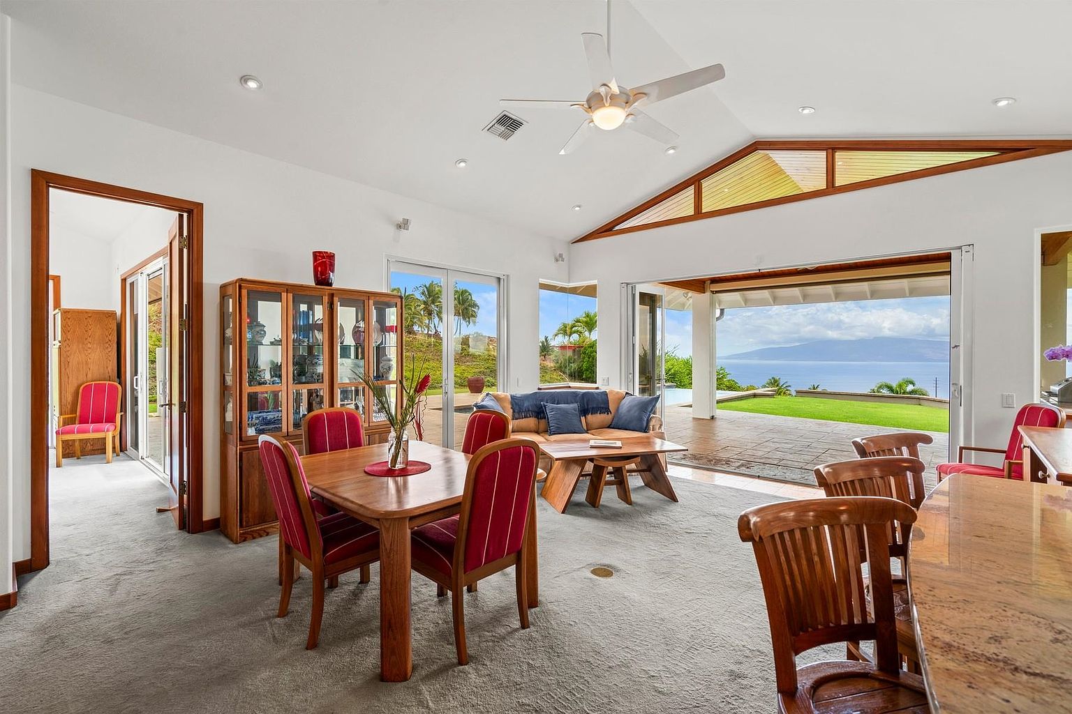 This is an interior shot of a living room that seamlessly blends indoor and outdoor living. The room features a dining area with a wooden table and red-cushioned chairs, a comfortable seating area with a sofa and coffee table, and large sliding glass doors that open to a patio with ocean views. The neutral color palette and natural materials create a relaxed and inviting atmosphere.