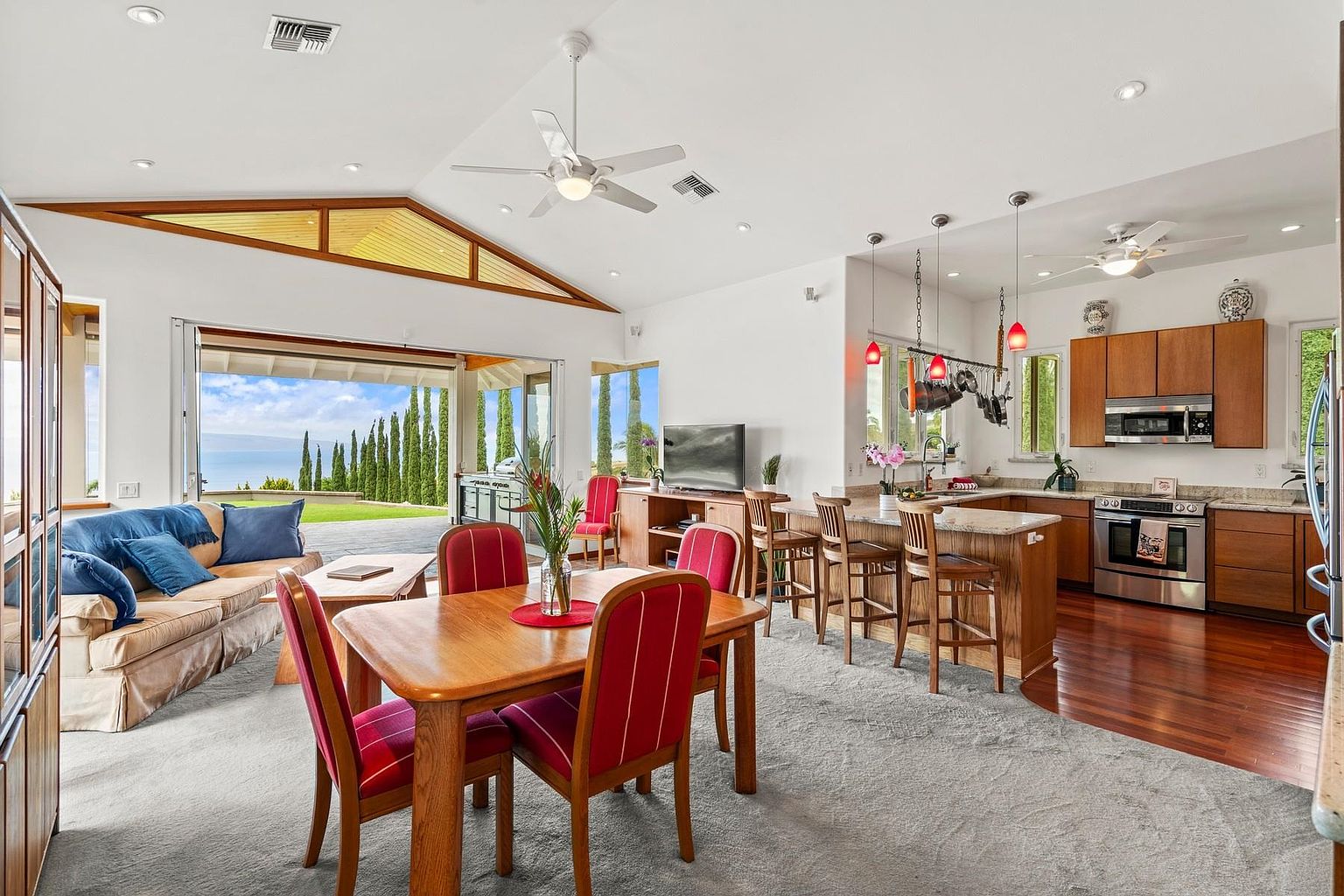 This is an interior shot showcasing an open-concept living space that seamlessly blends the living room, dining area, and kitchen. The living room features a comfortable sofa and a large opening to an outdoor view. The dining area has a wooden table with red chairs, and the kitchen is equipped with modern appliances and a breakfast bar. The overall impression is one of spaciousness and modern comfort.