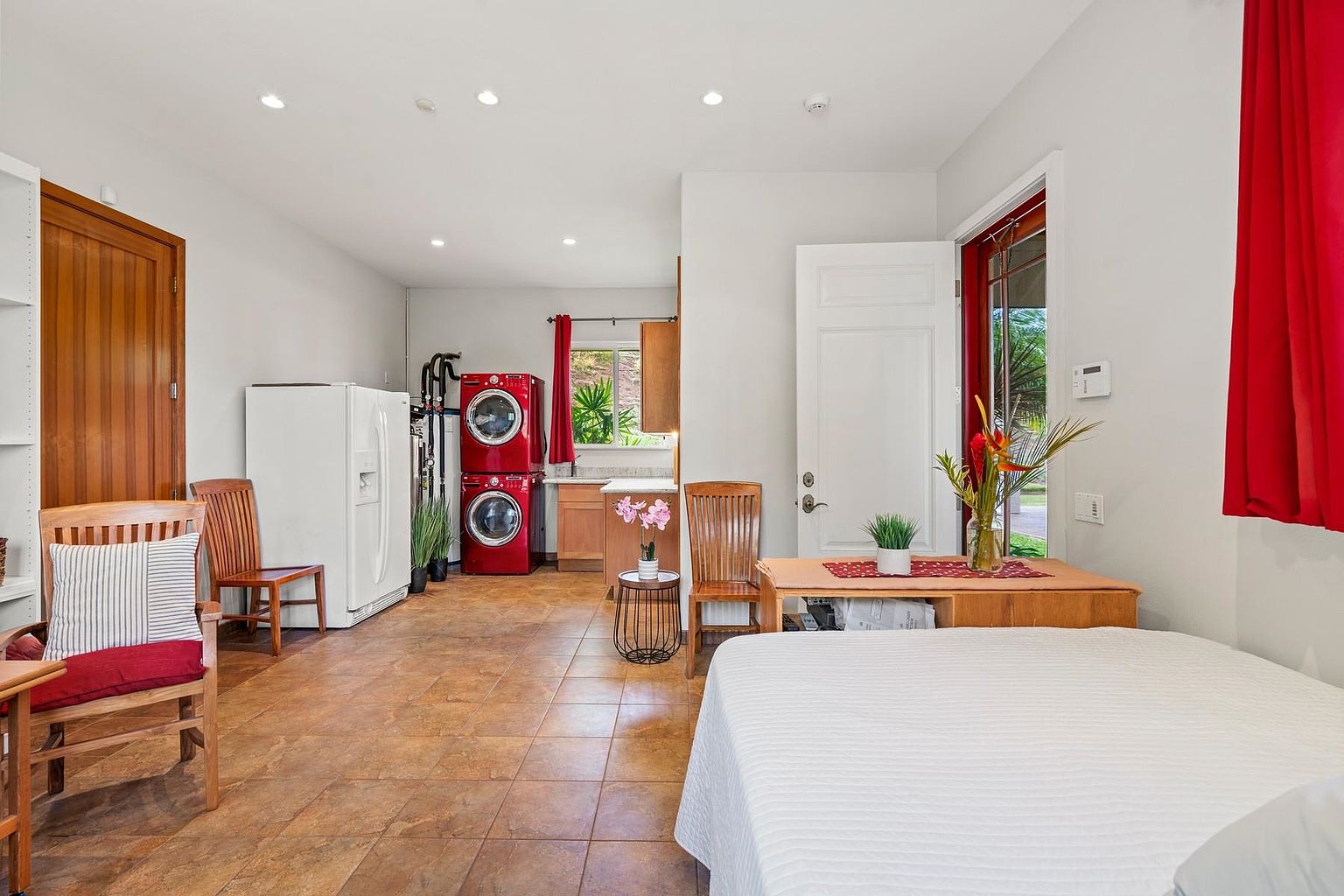This interior shot showcases a combined living space featuring a bedroom area with a bed covered in a white quilt. The room also includes a compact kitchen area with red stacked washer and dryer units, a refrigerator, and wooden cabinetry. The flooring is tiled, and the room is illuminated by recessed lighting, creating a functional and cozy atmosphere.