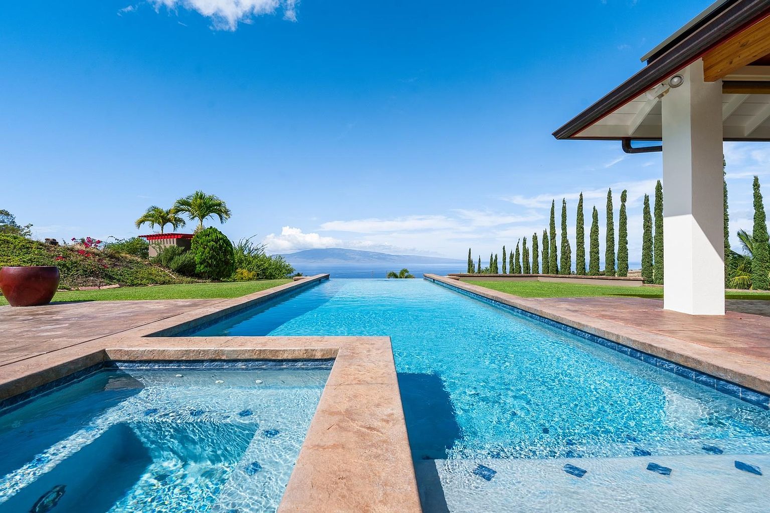 This image showcases a luxurious infinity pool and spa area with stunning ocean views. The pool's design seamlessly blends with the horizon, creating a sense of tranquility and openness. Lush landscaping and architectural details, such as the white pillar and manicured trees, enhance the property's appeal, suggesting a high-end, resort-like atmosphere.