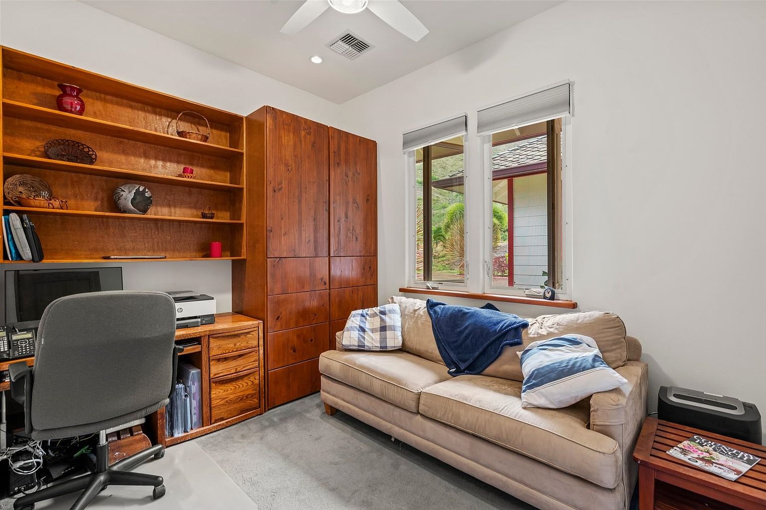 This is an interior shot of a home office or study. The room features a built-in wooden bookshelf and cabinet, a desk area with a computer, and a comfortable sofa with pillows and a throw blanket. Natural light streams in through the windows, creating a warm and inviting atmosphere.