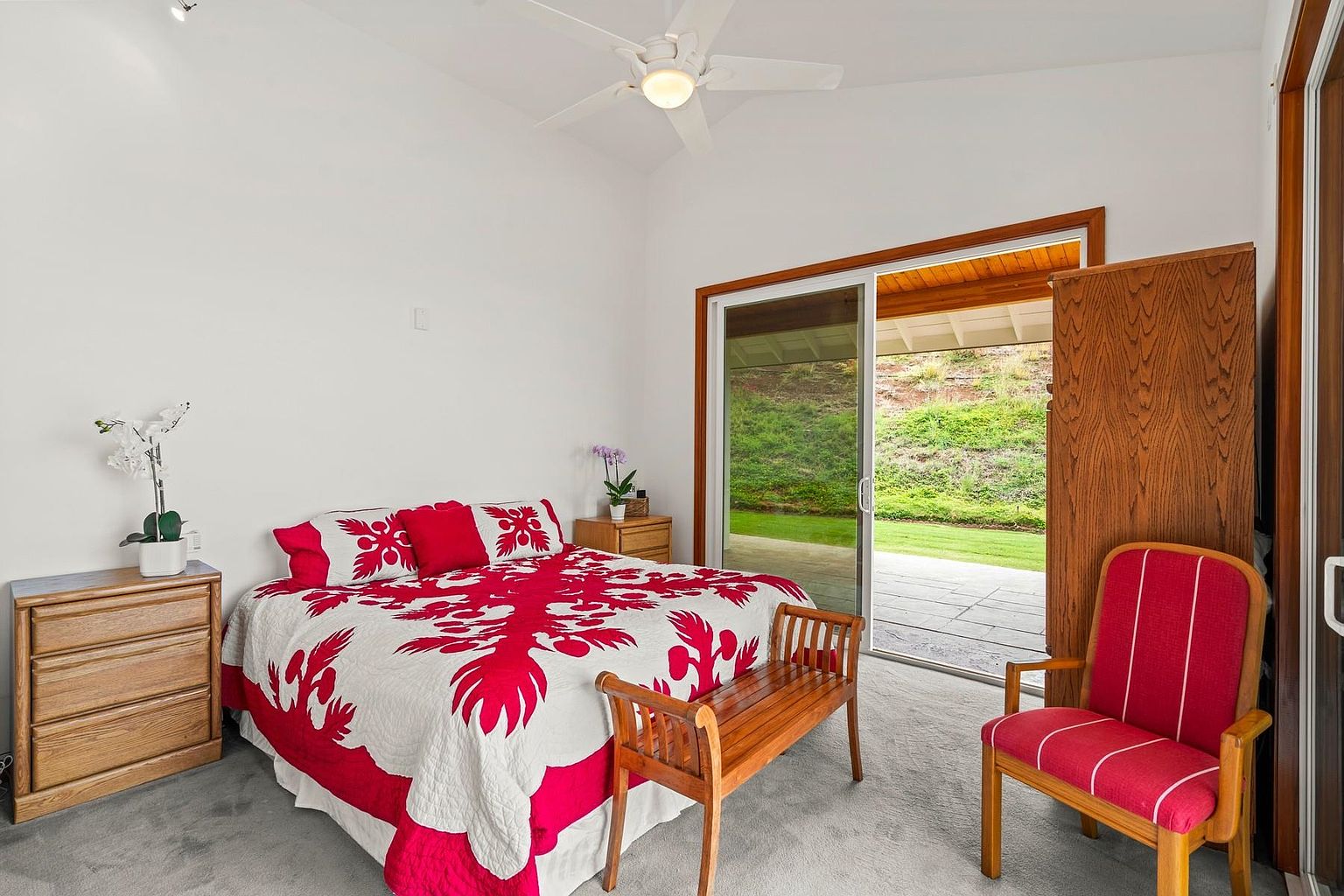 This is a bright and airy primary bedroom featuring a queen-sized bed with a red and white Hawaiian quilt. The room has white walls, a ceiling fan, and a sliding glass door that leads to an outdoor patio. A wooden dresser and a red upholstered chair add warmth and character to the space.