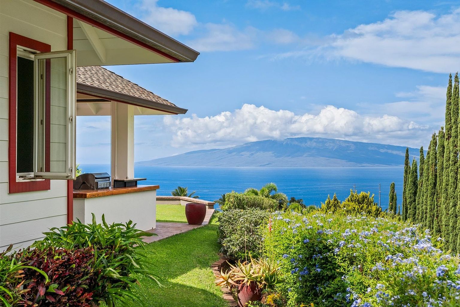 This image showcases a beautifully landscaped yard with lush greenery and vibrant blue flowers, offering a serene outdoor space. A partial view of the house reveals a covered patio with a built-in grill, suggesting an ideal setting for outdoor entertaining. The backdrop features a stunning ocean view with a distant island, enhancing the property's appeal and creating a sense of tranquility.