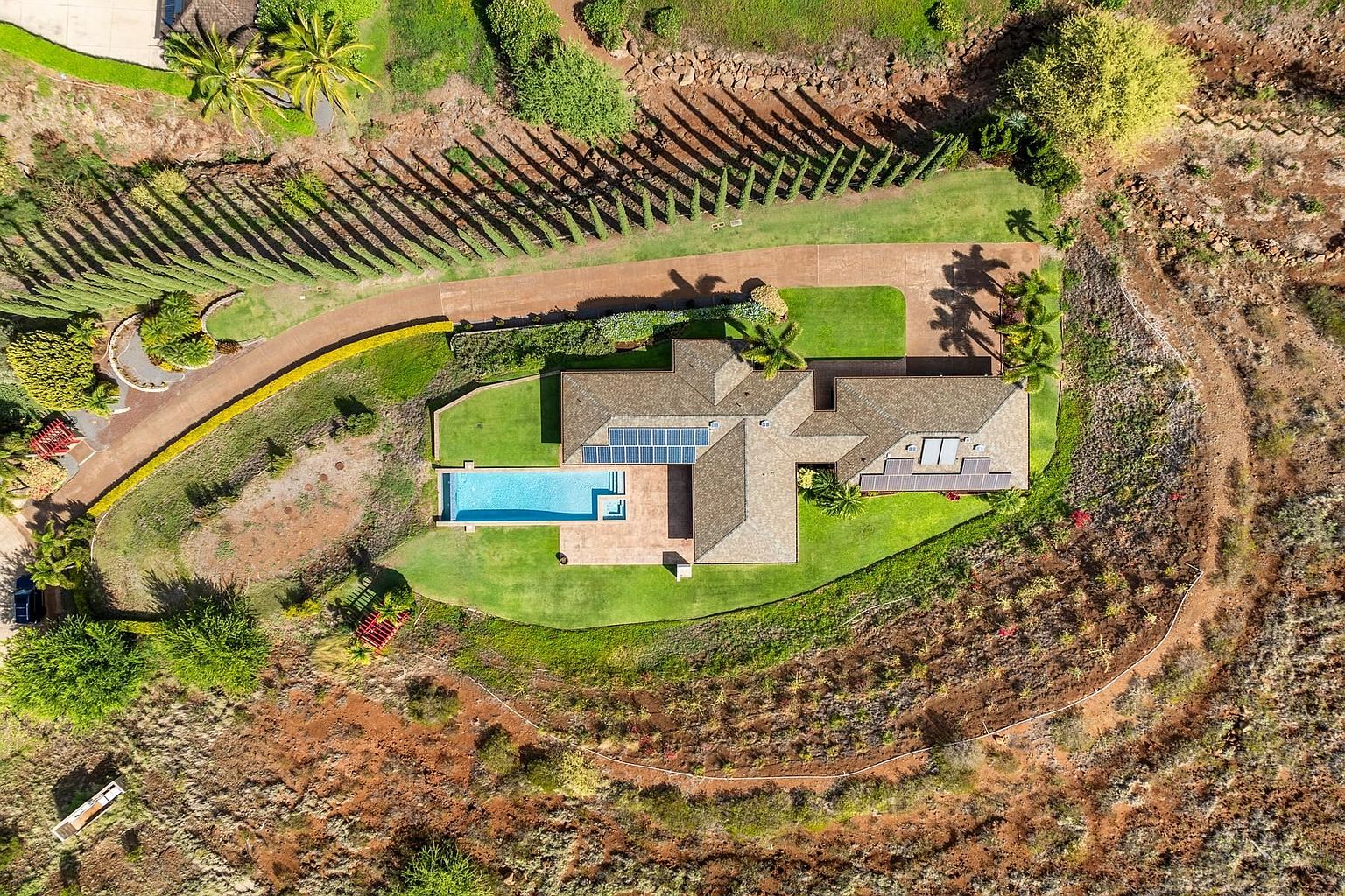 This aerial shot showcases a luxurious property featuring a large house with a swimming pool, surrounded by lush green lawns and mature landscaping. The house has a complex roofline with solar panels, and a long driveway leads to the property. The surrounding terrain is a mix of manicured gardens and natural, arid landscape, creating a sense of privacy and exclusivity.