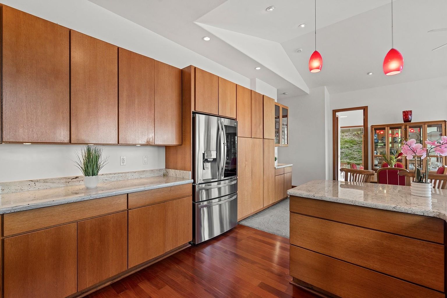 This is a well-lit kitchen featuring wooden cabinetry, stainless steel appliances, and granite countertops. The kitchen island provides additional workspace, and the hardwood floors add warmth to the space. Red pendant lights hang above the island, adding a pop of color.