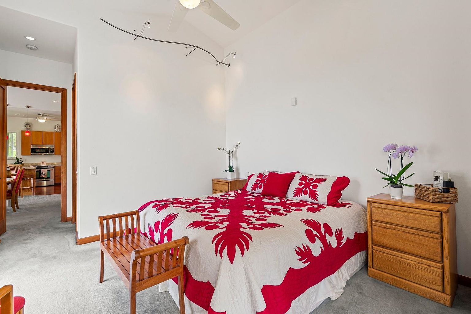 This is a cozy guest bedroom featuring a bed with a red and white Hawaiian quilt, wooden nightstands, and a matching wooden bench at the foot of the bed. The walls are painted white, and the room is well-lit, creating a clean and inviting atmosphere. A glimpse into the kitchen can be seen through an open doorway, adding depth to the scene.