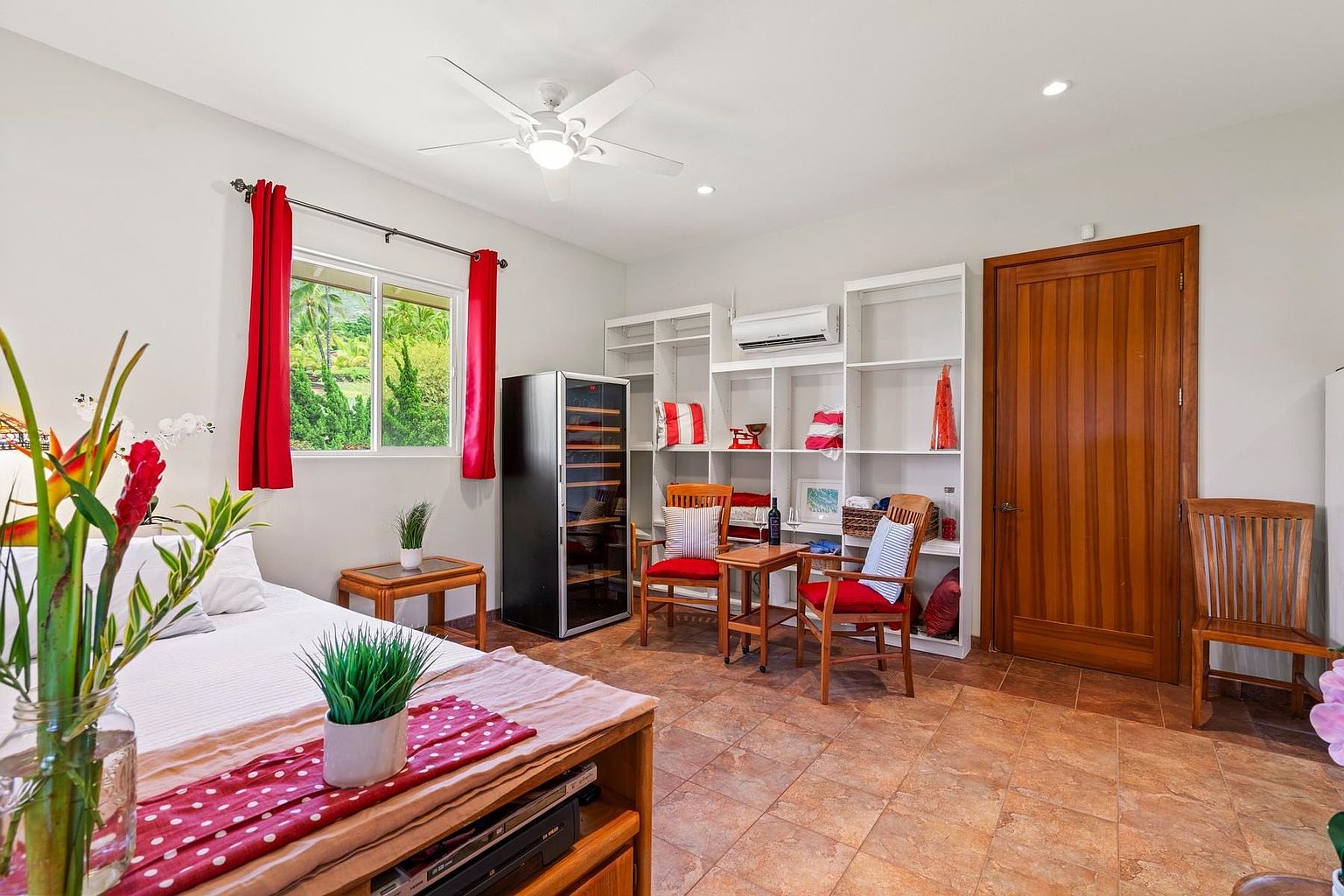 This is a cozy bedroom featuring a bed with red accents, a window with red curtains, and a seating area with two wooden chairs and a small table. Built-in white shelving provides storage, and a wine cooler adds a touch of luxury. The room has a warm and inviting atmosphere with tiled floors.