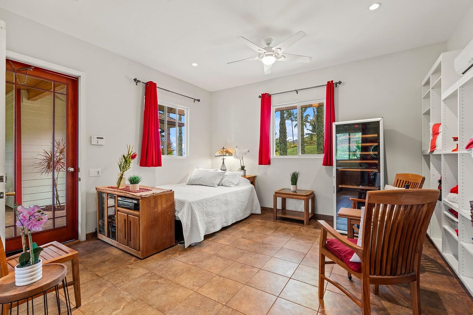 This is a cozy guest bedroom featuring a comfortable bed with white linens, flanked by wooden nightstands and windows adorned with red curtains. A wooden cabinet with glass doors sits near the entrance, while a wine cooler and a wooden chair add to the room's functionality and charm. The room is well-lit with recessed lighting and natural light, creating a welcoming atmosphere.