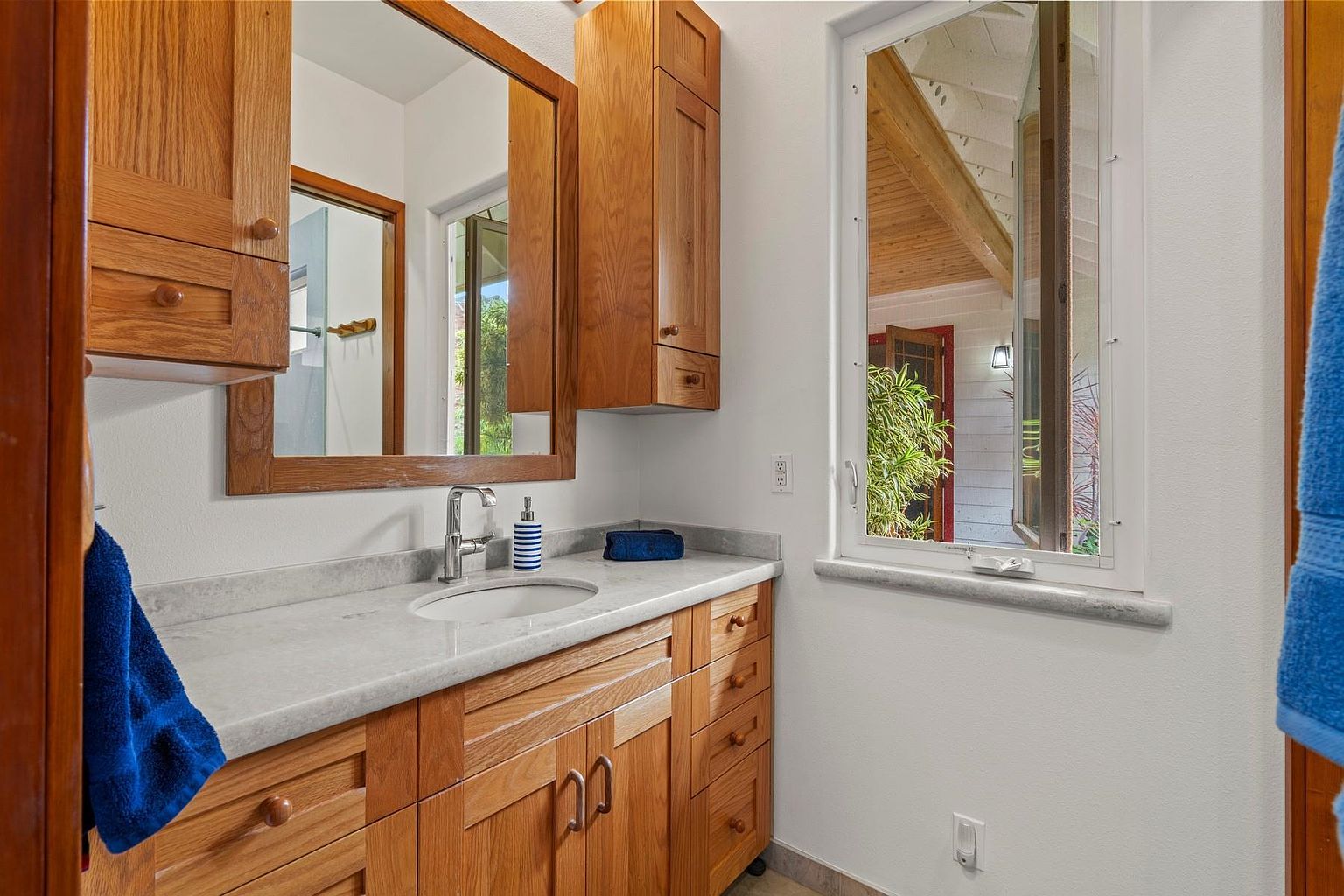 This is a well-lit bathroom featuring light wood cabinetry, a marble countertop, and a rectangular mirror. A window provides natural light and a view to the outside. The bathroom has a clean and functional design, suitable for a guest or secondary bathroom.