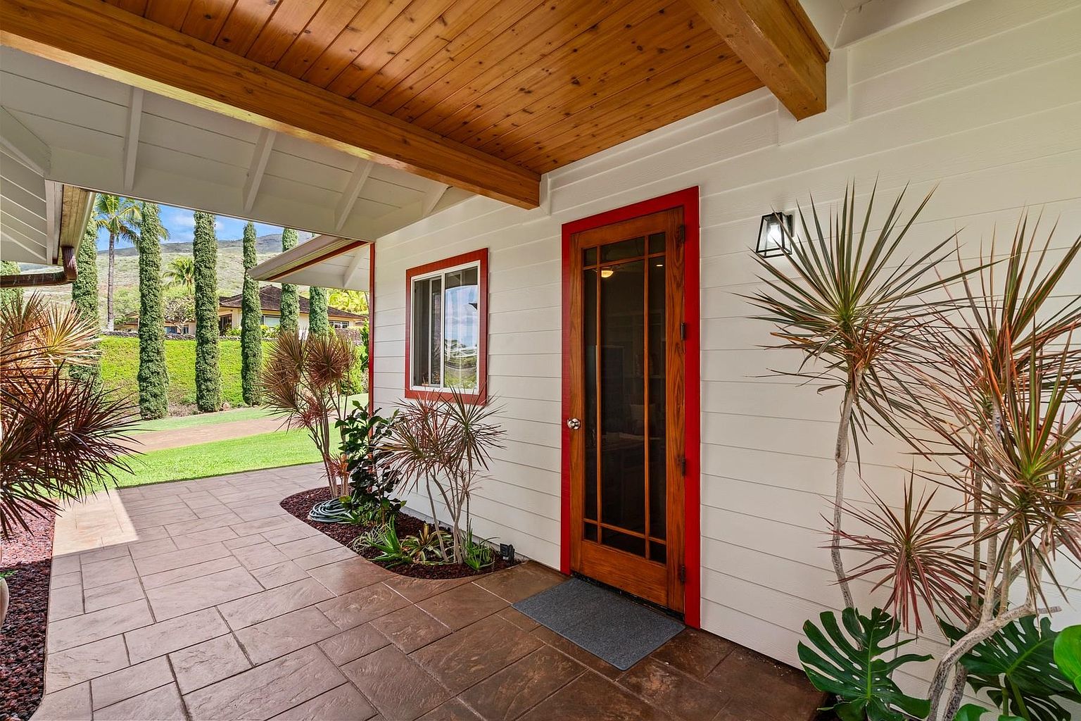 This image showcases an inviting entryway to a home, featuring a wooden door with red trim, a small window, and a covered porch area with a wooden ceiling. The exterior is painted white, and the landscaping includes various plants and trees, adding to the curb appeal. The walkway is paved with brown stone tiles, creating a warm and welcoming entrance.