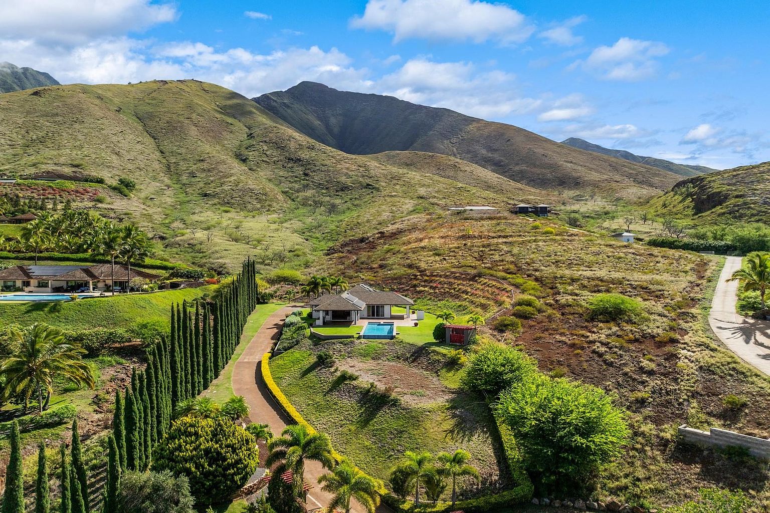 This aerial view showcases a luxurious property nestled in a mountainous landscape. The house features a modern design with a private pool, surrounded by lush greenery and a well-maintained lawn. A winding driveway leads to the residence, emphasizing its secluded and exclusive setting, with a cinematic perspective.