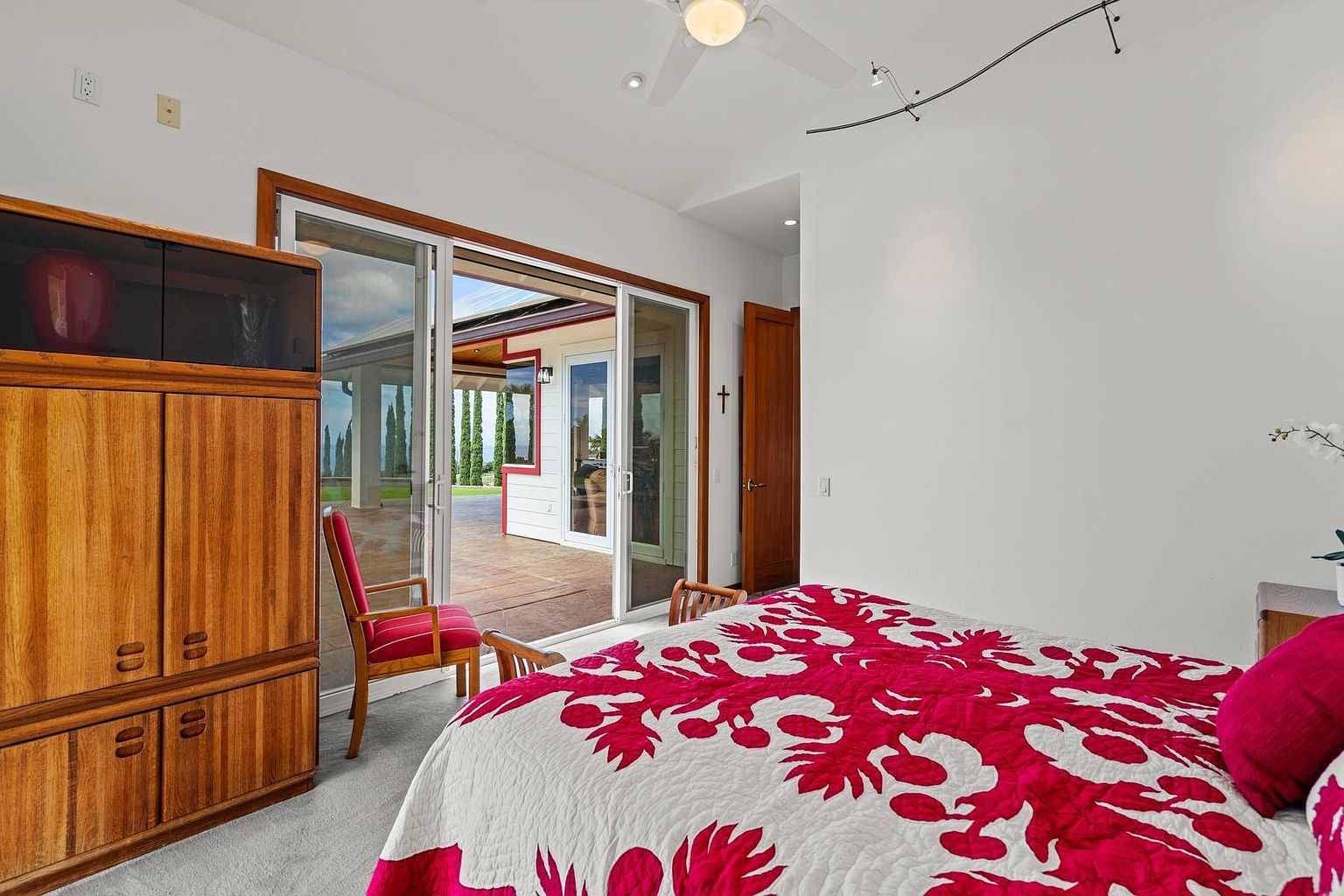 This primary bedroom features a bright and airy atmosphere with a large sliding glass door leading to an outdoor patio. The room is furnished with a wooden armoire and a bed adorned with a vibrant red and white quilt. The walls are painted a clean white, enhancing the sense of spaciousness and light.