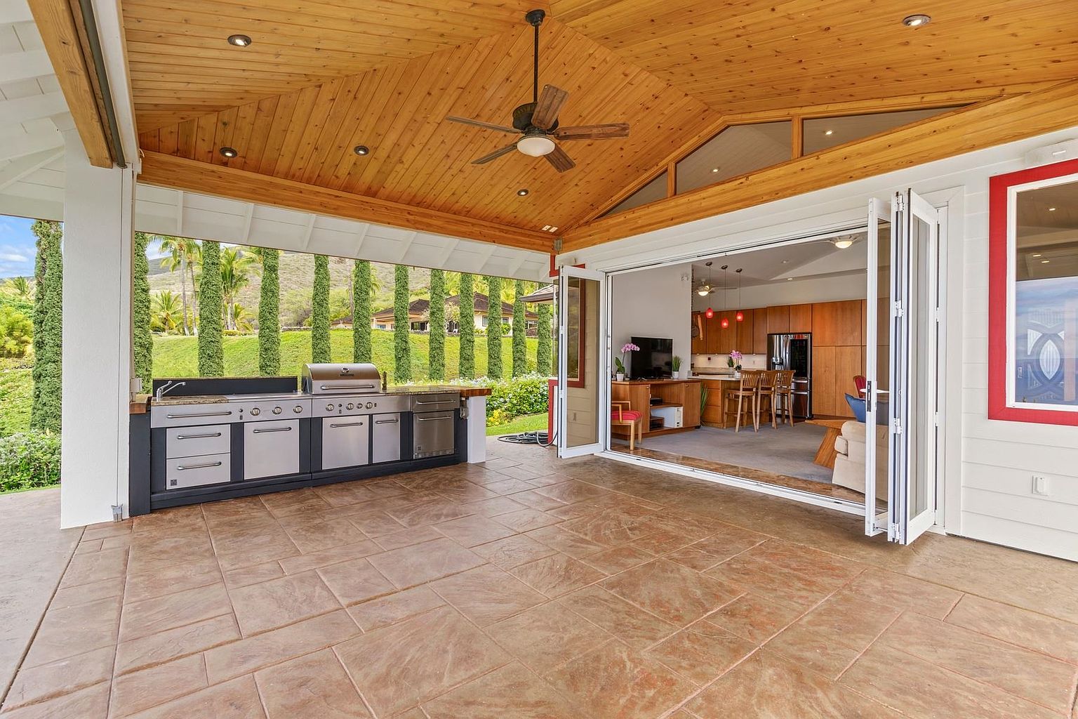 This image showcases a spacious outdoor patio area seamlessly connected to the interior through large, folding glass doors. The patio features a built-in outdoor kitchen with stainless steel appliances, a tiled floor, and a wooden ceiling with recessed lighting and a ceiling fan. The open design provides an inviting transition between indoor and outdoor living spaces, with a view of a well-maintained lawn and landscaping.