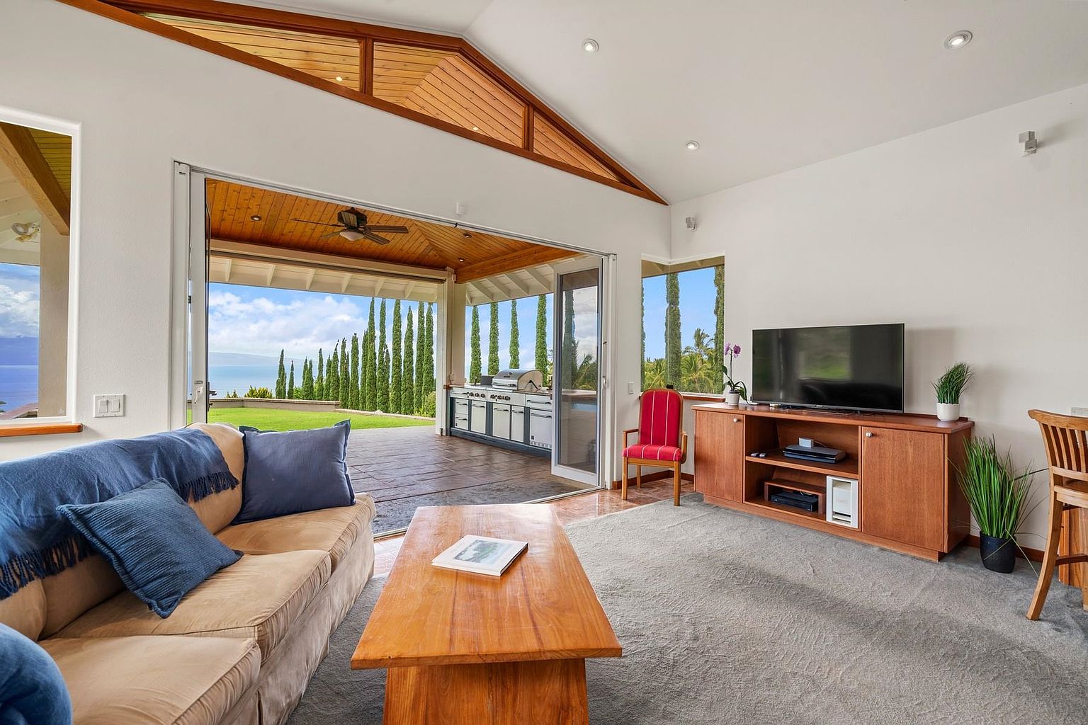 This is an interior shot of a living room that seamlessly blends indoor and outdoor living. The room features a comfortable beige sofa with blue cushions, a wooden coffee table, and a TV console with a flat-screen TV. Large sliding doors open to an outdoor patio with a built-in grill and a view of lush greenery and the ocean, creating a bright and airy atmosphere.