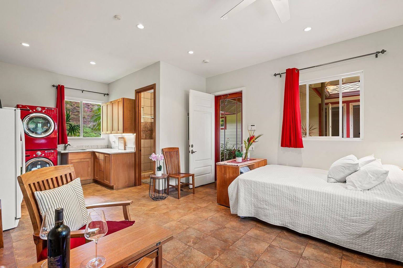 This is an interior shot of a guest bedroom that also functions as a living space. The room features a bed with white linens, a small desk area, and a kitchenette with red appliances. The flooring is tiled, and the room is well-lit with natural light coming through the windows, creating a cozy and functional space.