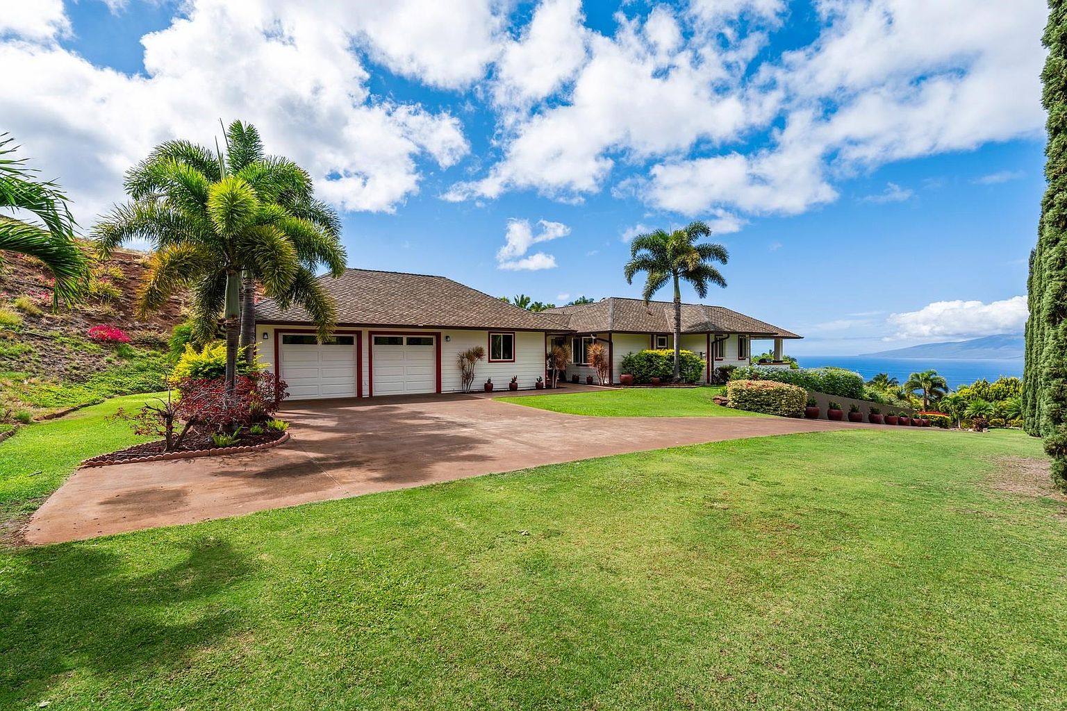 This is a front exterior view of a single-family home with a well-manicured lawn and mature landscaping. The house features a multi-car garage, a brown roof, and a long driveway leading up to the property. The ocean is visible in the background, suggesting a desirable location with scenic views.