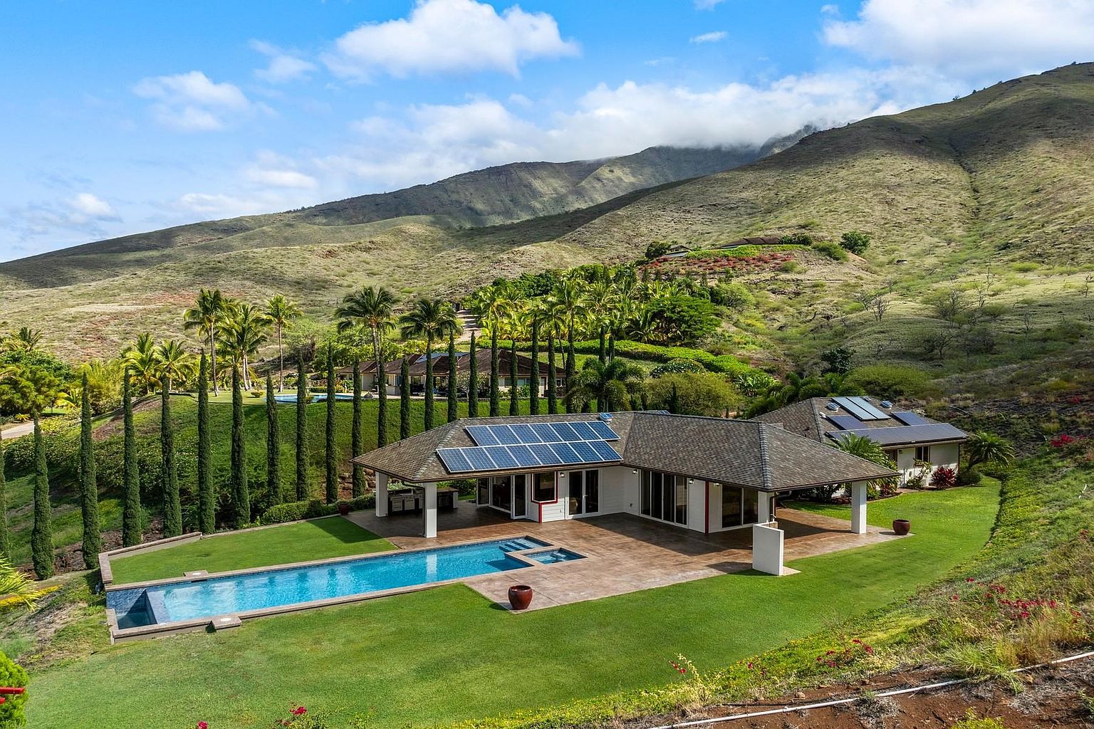 This aerial view showcases a luxurious property nestled against a scenic mountain backdrop. The house features solar panels, a spacious patio, and a stunning pool with a spa. Lush green lawns and tall trees surround the property, creating a private and serene atmosphere.