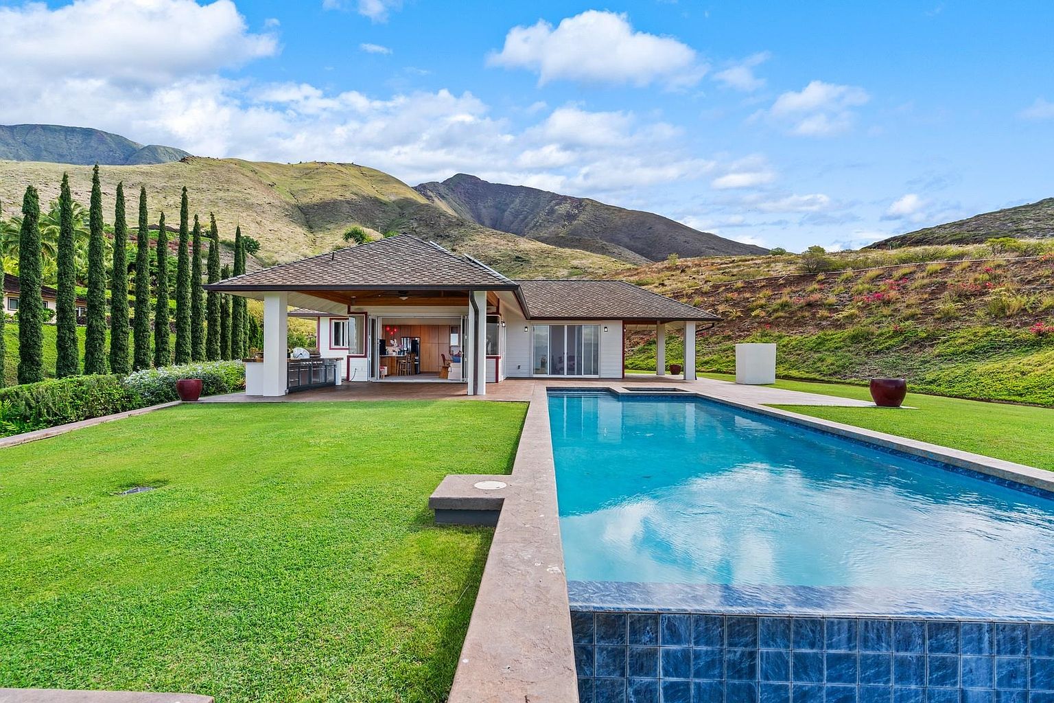 This image showcases a luxurious backyard featuring a stunning pool with blue tile accents, surrounded by a lush green lawn. The property includes a modern house with an open-air design, seamlessly blending indoor and outdoor living. Mountains in the background add to the serene and upscale ambiance, making it an ideal setting for relaxation and entertainment.
