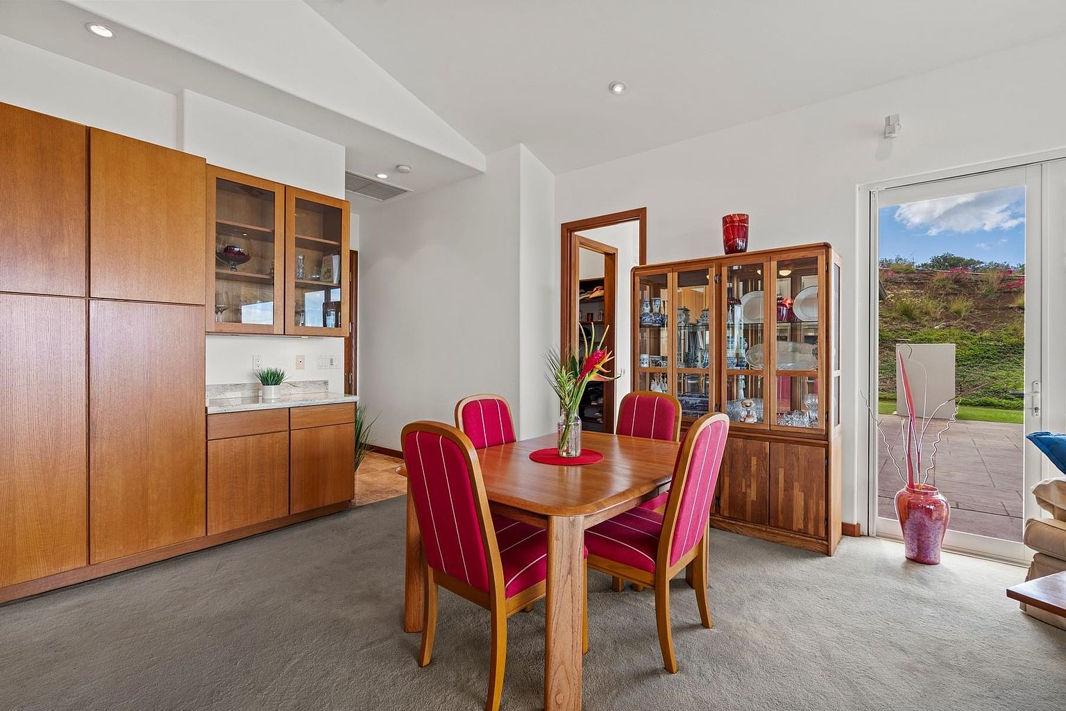This dining room features a wooden table with four chairs upholstered in red and white stripes, creating a warm and inviting atmosphere. A built-in wooden cabinet and a separate display cabinet showcase decorative items, adding character to the space. Natural light streams in through a sliding glass door, offering a glimpse of the outdoor landscape.