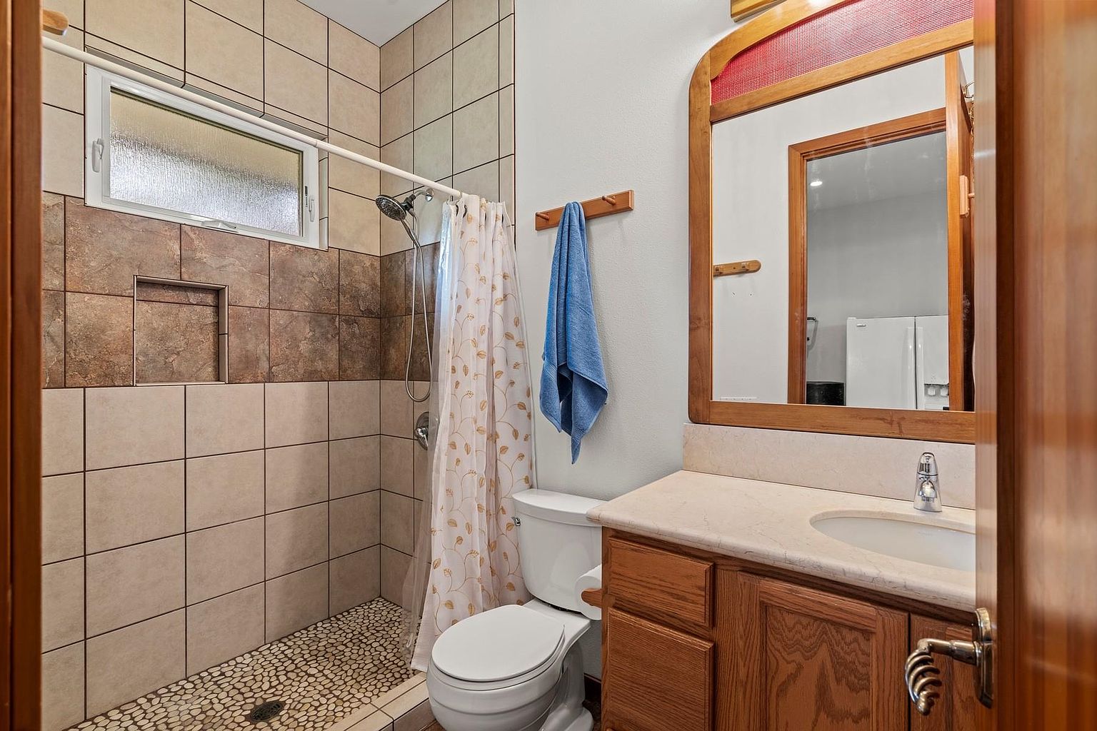 This is a bathroom featuring a tiled shower with a pebble floor and a shower curtain. The vanity has a wooden cabinet and a light-colored countertop with an oval sink. A wooden-framed mirror hangs above the vanity, and a blue towel is on a wooden rack next to the toilet.