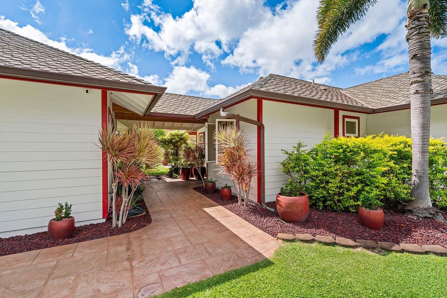 This image showcases the entryway of a well-maintained home, featuring a paved walkway leading between two sections of the house. The exterior is painted white with red trim, complemented by lush landscaping including potted plants and decorative rock. The overall impression is inviting and well-cared for, highlighting the property's curb appeal.