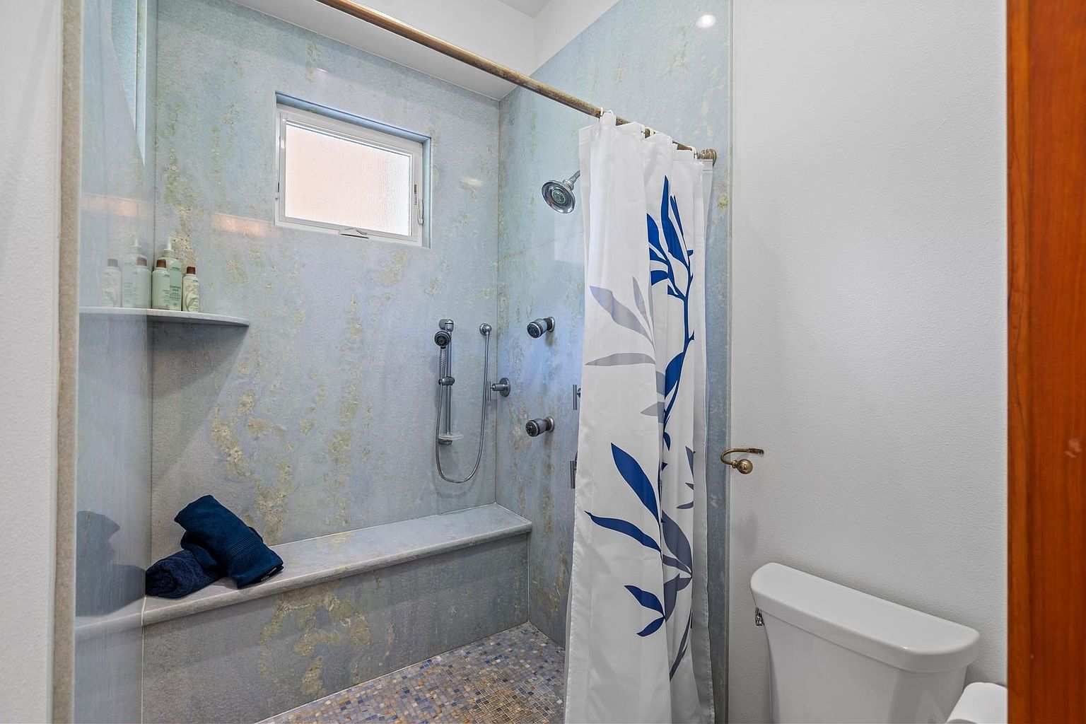 This is a bathroom featuring a walk-in shower with a built-in bench and a shower curtain with a blue leaf pattern. The shower walls are tiled in a light blue marble-like material, and there's a small window above the shower controls. A toilet is visible next to the shower, and the overall impression is clean and functional.
