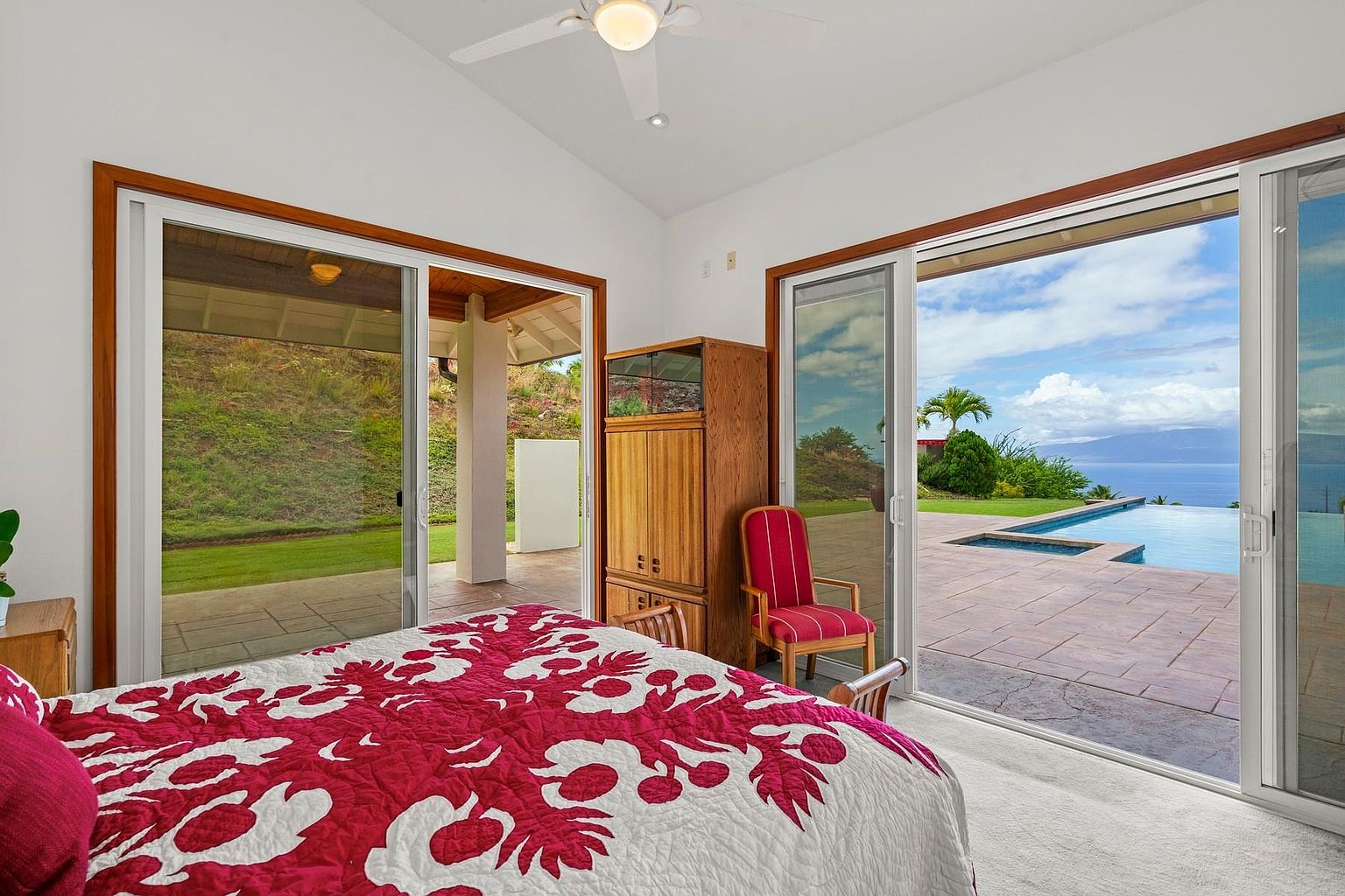 This is a primary bedroom featuring a large bed with a red and white patterned quilt. Sliding glass doors open to a patio with a pool and ocean view, creating a seamless indoor-outdoor living experience. A wooden armoire and a red chair add to the room's decor.