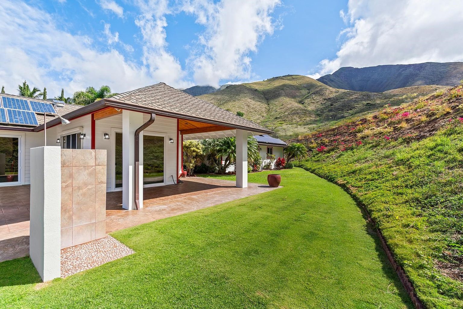 This image showcases a well-maintained yard with lush green grass and a landscaped hillside. A covered patio extends from the house, featuring a tiled outdoor shower. The backdrop includes a scenic mountain view, enhancing the property's appeal.