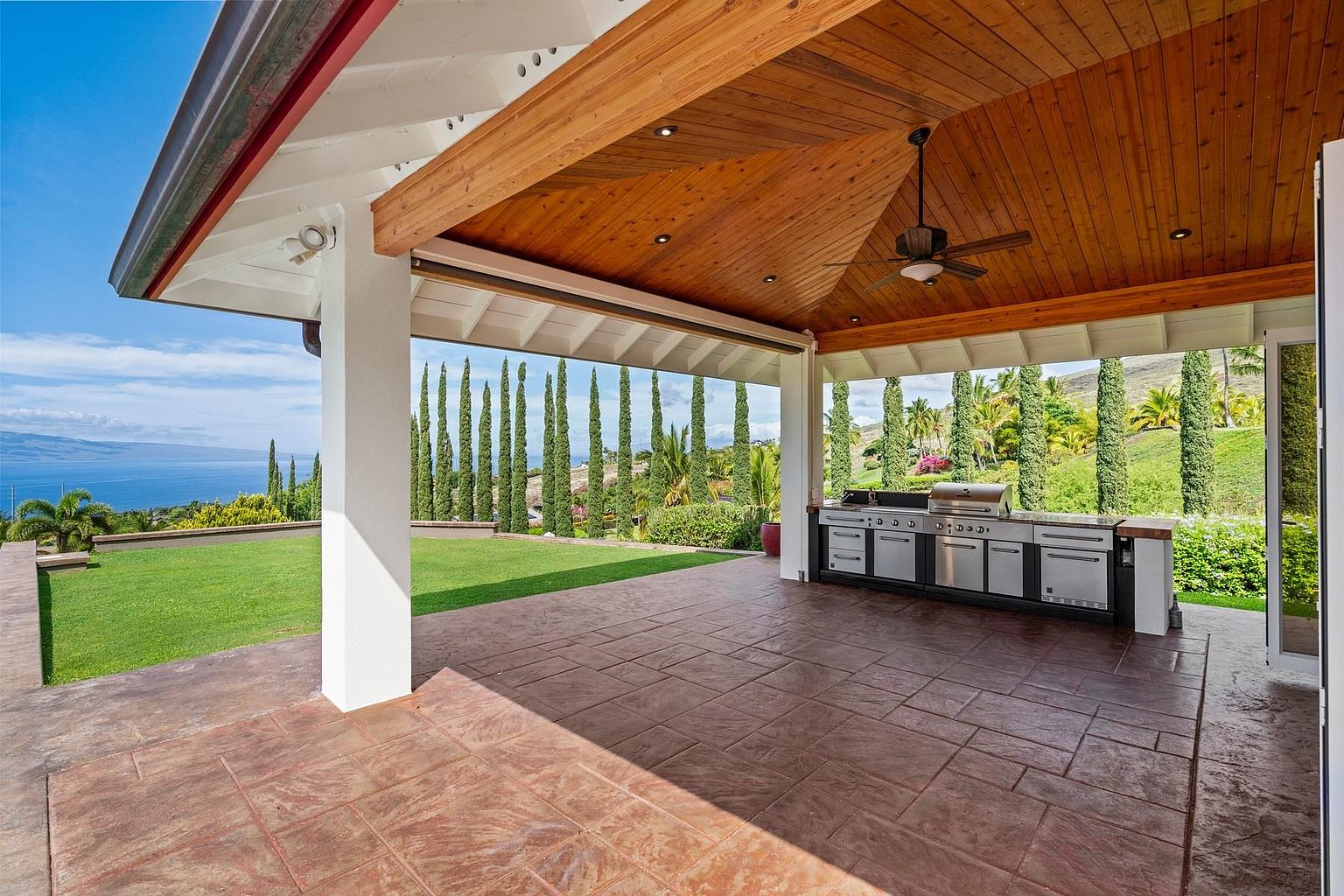 This image showcases a covered outdoor patio area, featuring a built-in stainless steel grill and countertop setup. The patio has a textured tile floor and is framed by white columns and a wooden ceiling, offering a blend of functionality and style. The view extends to a well-manicured lawn, tall cypress trees, and a distant ocean view, enhancing the property's appeal for outdoor living and entertaining.
