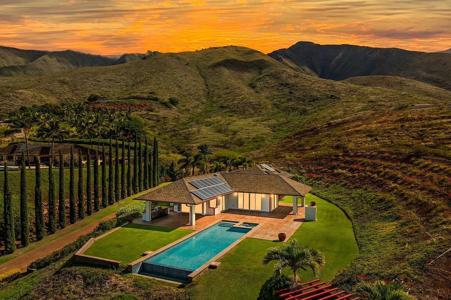This aerial view showcases a luxurious home nestled in a mountainous landscape, featuring a modern design with solar panels, a private pool, and meticulously manicured lawns. The property is surrounded by lush greenery and tall, slender trees, creating a serene and secluded atmosphere. The warm sunset adds a touch of drama and highlights the architectural details of the house.