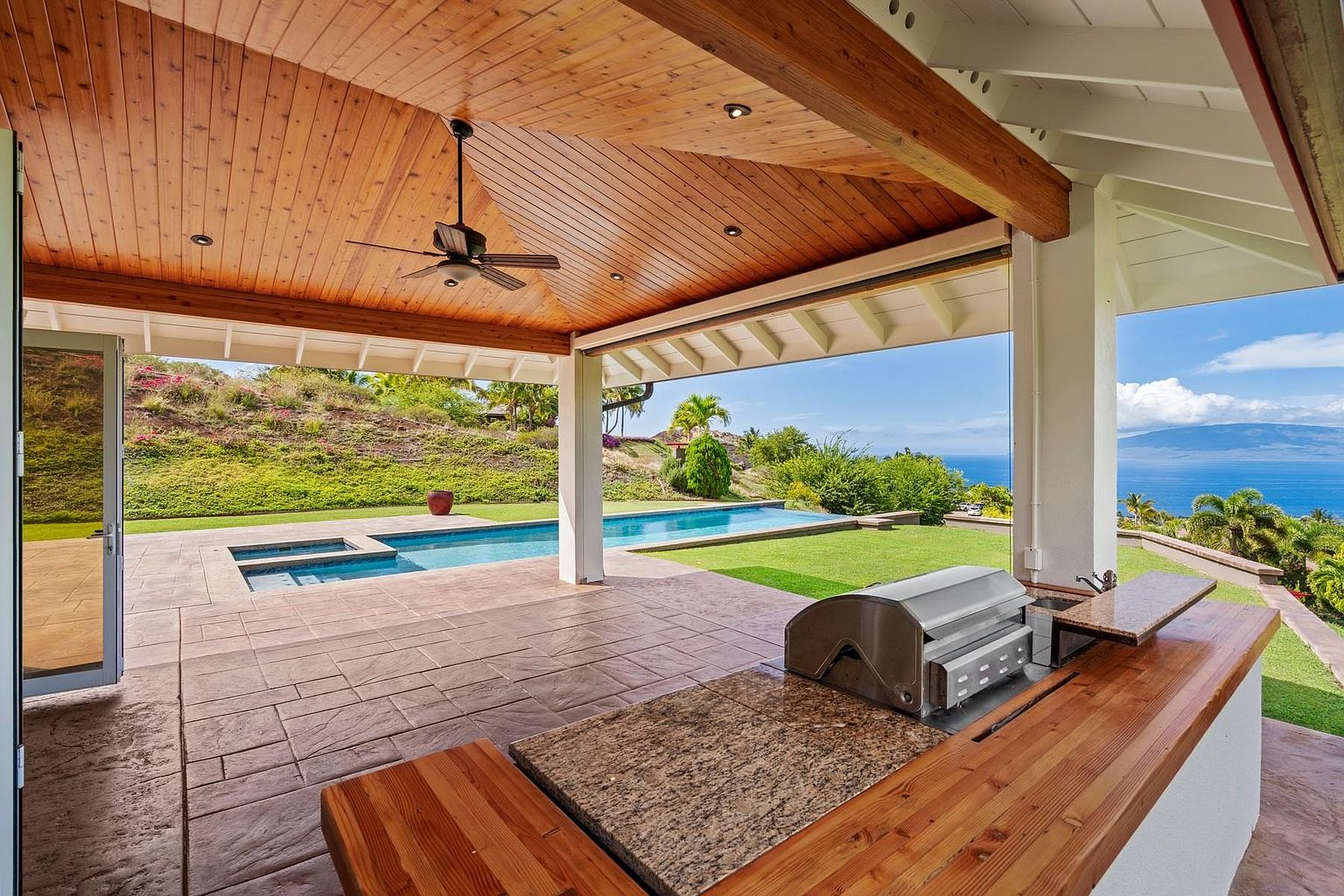 This image showcases a luxurious outdoor patio area with a built-in grill and countertop, overlooking a stunning ocean view. The patio features a wooden ceiling with a ceiling fan, stone flooring, and a nearby swimming pool. The overall impression is one of high-end outdoor living and relaxation.