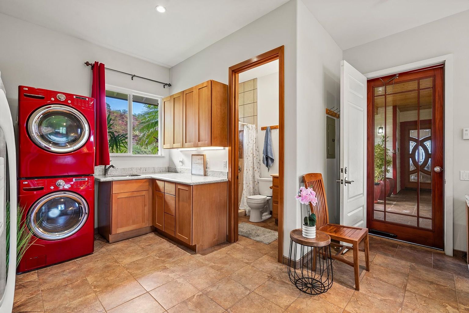 This interior shot showcases a laundry room with a vibrant red stacked washer and dryer set. The room features wooden cabinetry with light countertops, a window providing natural light, and a doorway leading to a bathroom. The tile flooring adds a warm tone to the space, and a decorative chair and table are placed near the entrance.