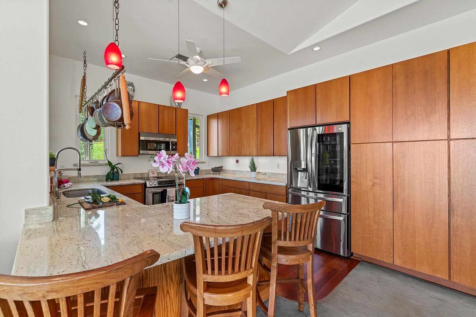 This is a well-lit kitchen featuring wooden cabinetry and stainless steel appliances, including a large refrigerator. A granite countertop island with bar seating is in the foreground, adorned with a vase of pink orchids. The kitchen also has a pot rack hanging above the sink area, adding a touch of rustic charm.