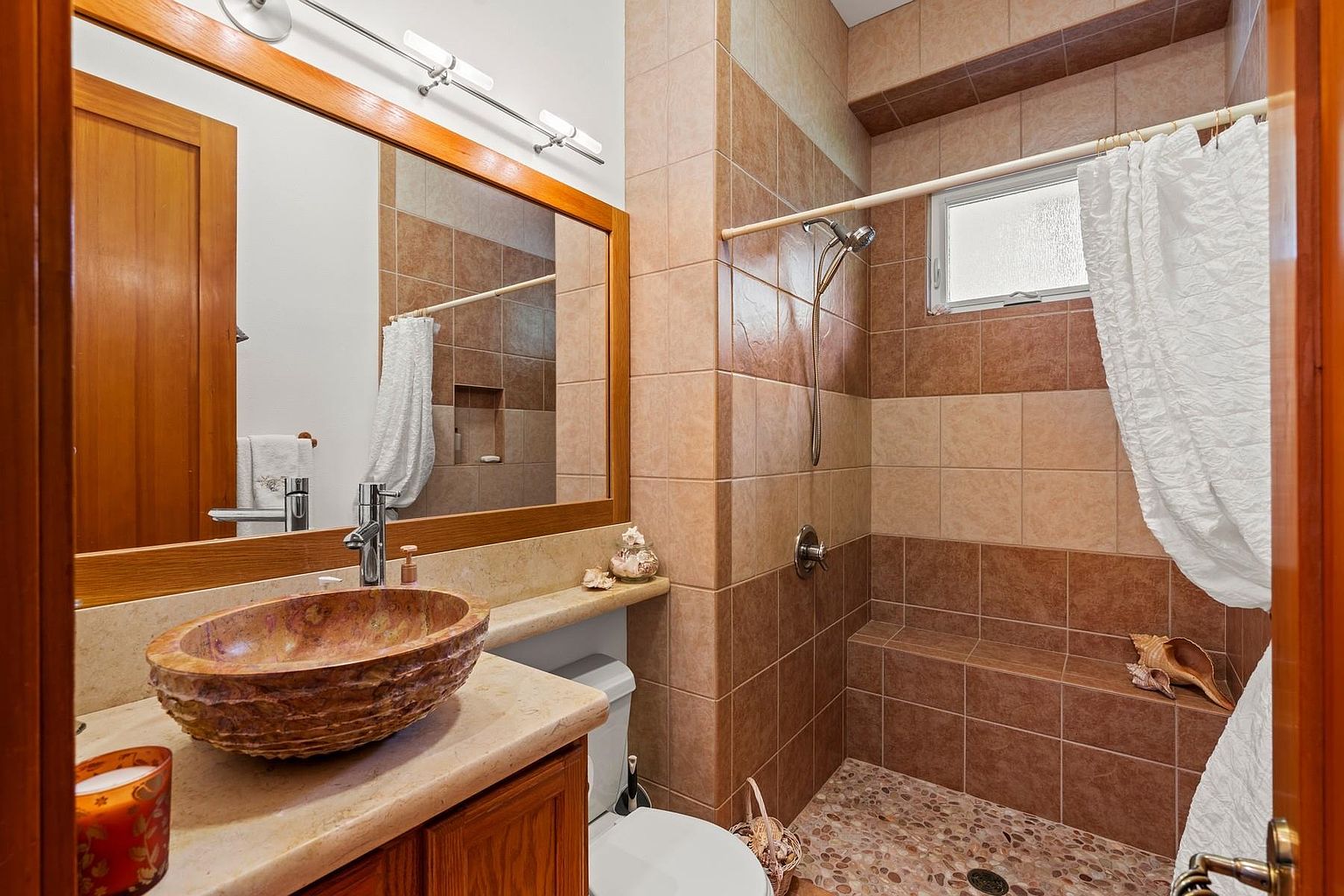 This is a bathroom featuring a unique stone vessel sink on a wooden vanity with a light-colored countertop. The shower area is tiled in alternating shades of brown and beige, with a pebble floor and a built-in bench. A framed mirror hangs above the sink, reflecting the bathroom's details and creating a warm, inviting atmosphere.
