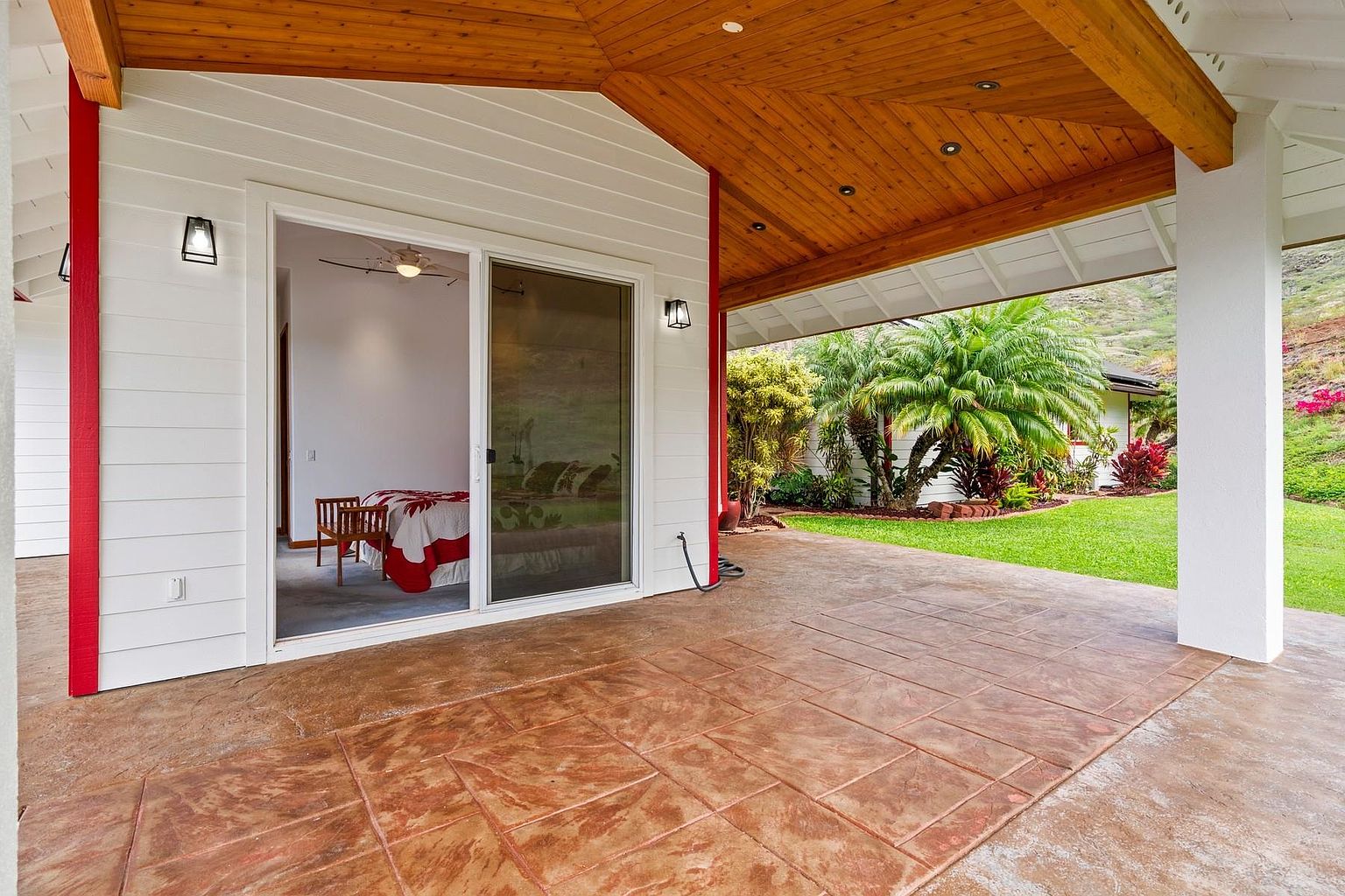 This image showcases a covered patio area with a decorative concrete tile floor, seamlessly blending indoor and outdoor living. The patio connects to an interior room through a sliding glass door, offering easy access and a view of the bedroom. The space is framed by wooden beams and a white support column, with a glimpse of a well-maintained garden and lawn beyond, creating an inviting and relaxing atmosphere.