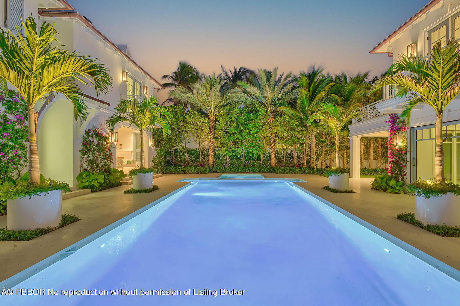 This image showcases a luxurious backyard pool area at dusk. The rectangular pool, illuminated with a soft blue glow, is the focal point, surrounded by a meticulously landscaped courtyard with palm trees in planters and lush greenery. The architecture of the surrounding buildings features a clean, white facade, enhancing the overall elegant and serene atmosphere.