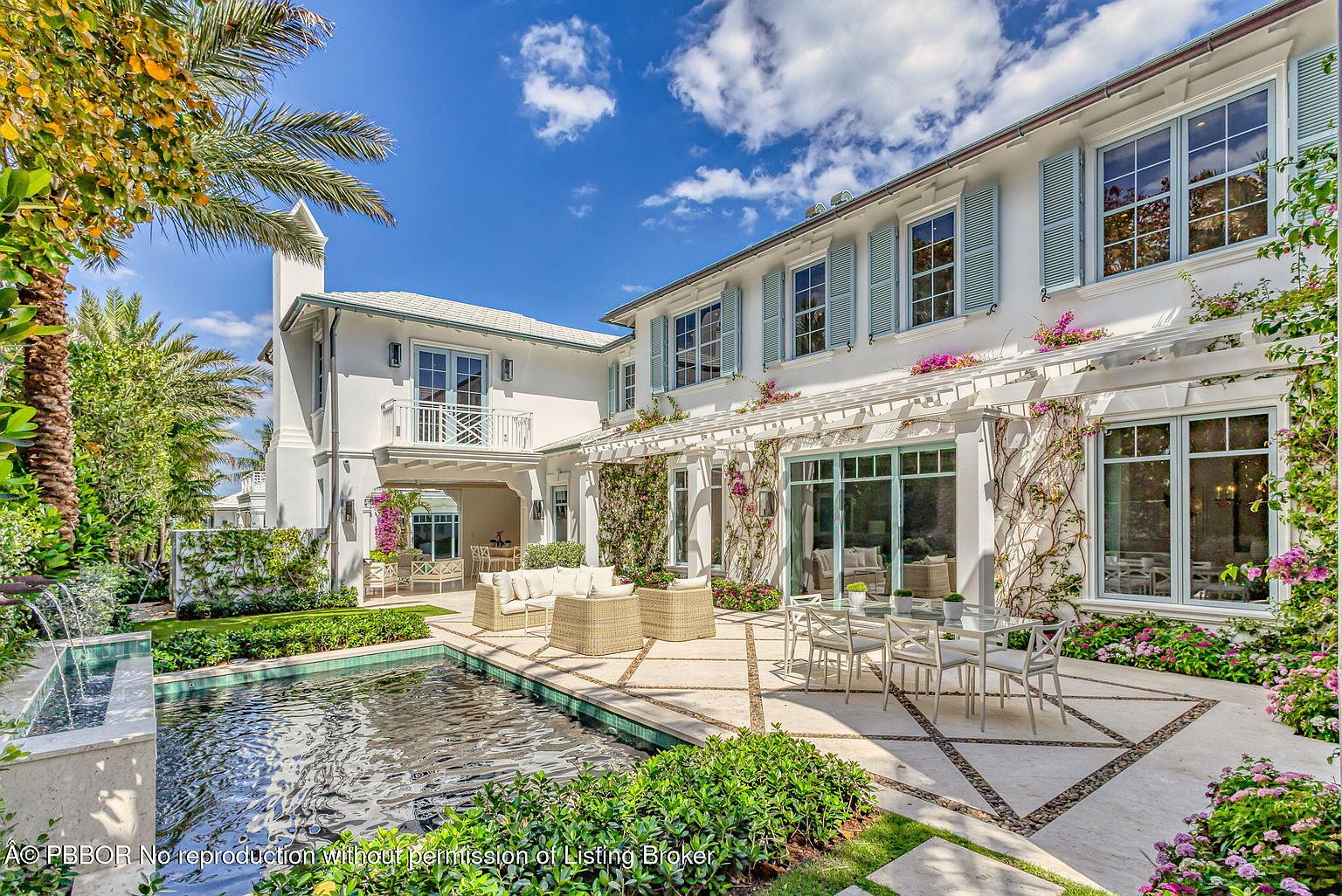 This image showcases a luxurious outdoor patio area, featuring elegant patio furniture, a dining set, and a tranquil pool. The architecture is characterized by a white facade with light blue shutters, complemented by climbing vines and vibrant floral arrangements. The overall impression is one of sophisticated outdoor living and relaxation.