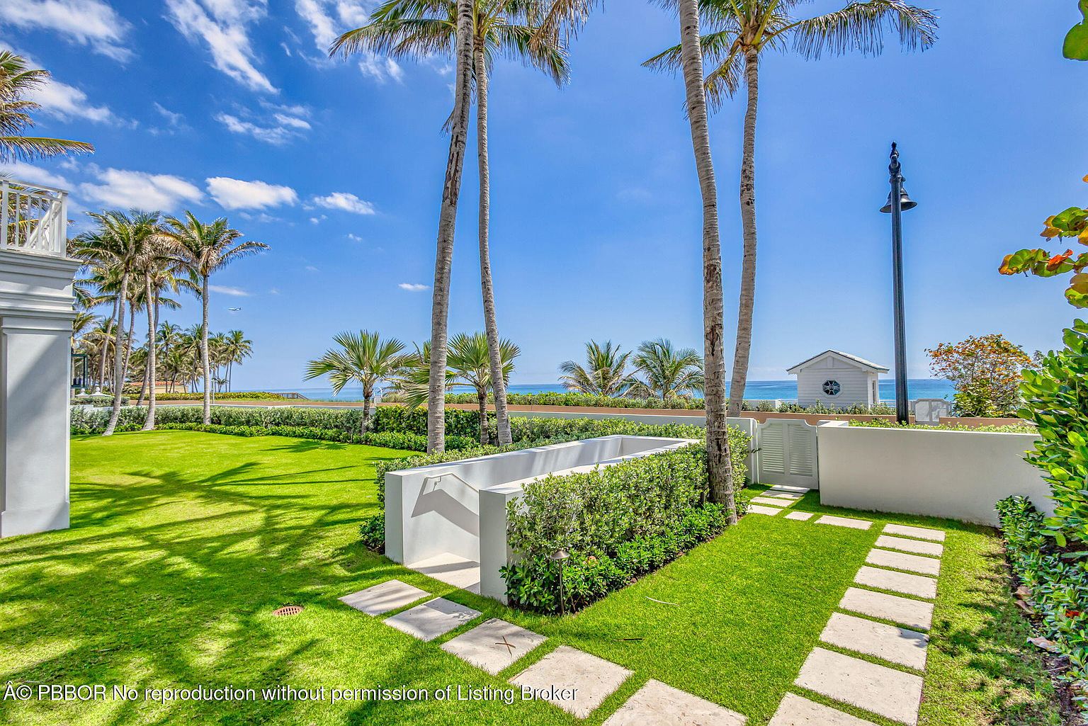This image showcases a beautifully landscaped yard with lush green grass, palm trees, and a glimpse of the ocean in the background. A white structure, possibly a pool feature or decorative element, is surrounded by manicured hedges. Stone pavers create a pathway through the grass, leading towards a white gate and a small building near the beach, creating an inviting and serene outdoor space.