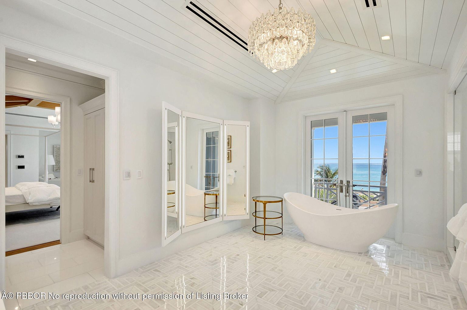 This is a luxurious primary bathroom featuring a freestanding soaking tub positioned in front of French doors that open to a balcony with ocean views. The room is decorated in a bright, all-white color scheme with a crystal chandelier, a large folding mirror, and a patterned marble floor. The overall impression is one of elegance and relaxation.