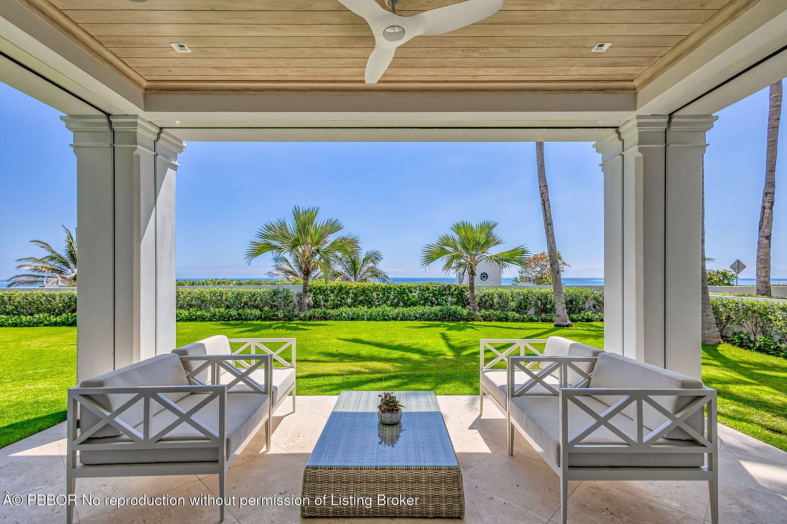 This image showcases a luxurious outdoor patio or balcony area with a stunning view of the ocean and lush greenery. The space is furnished with elegant, light-colored sofas and a stylish coffee table, creating an inviting and relaxing atmosphere. The architectural details, including the white columns and wood-paneled ceiling with a ceiling fan, add to the overall sophistication and appeal of the property.