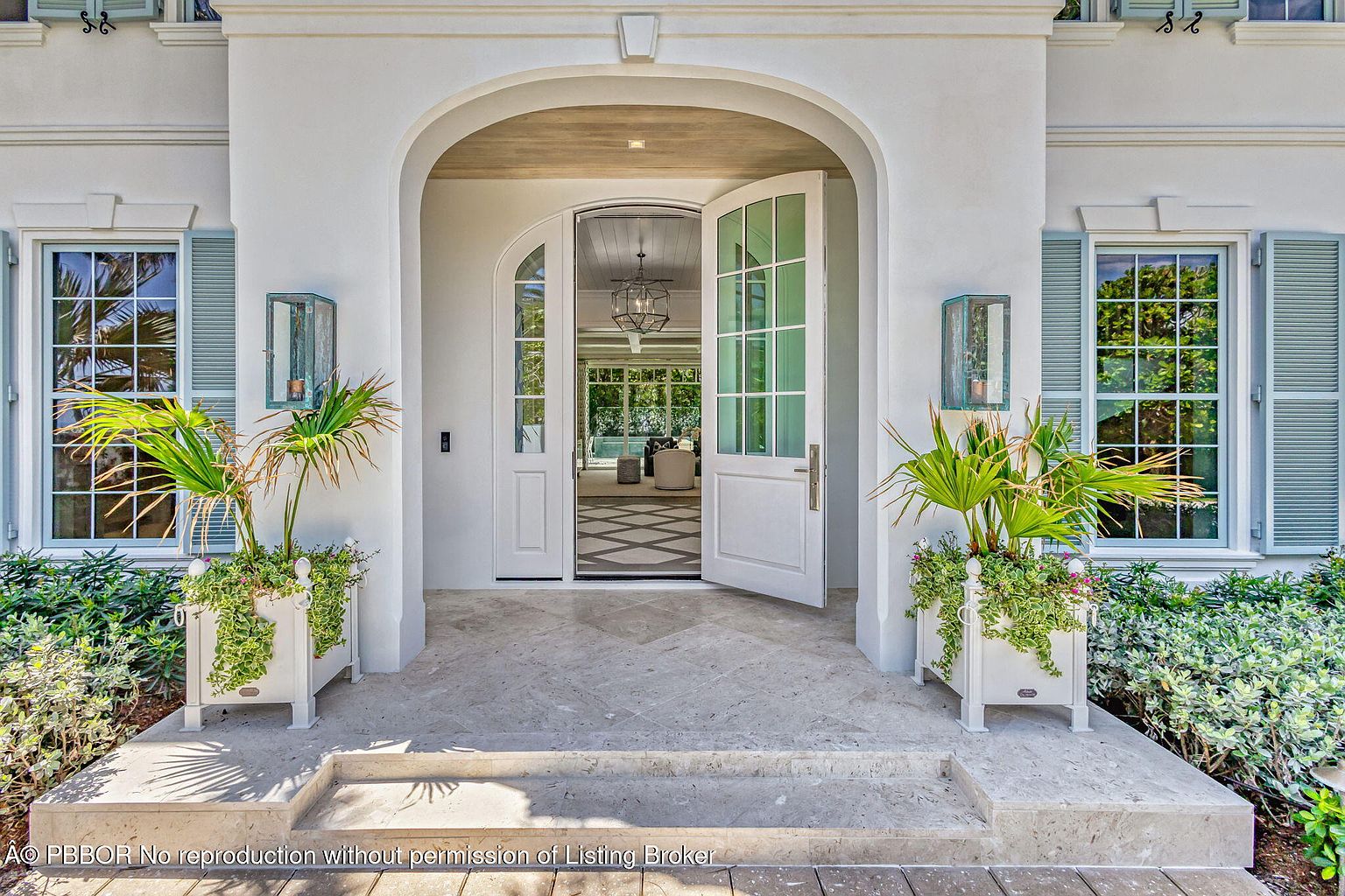 This is a welcoming entryway to a luxurious home, featuring a grand arched doorway with double doors that are partially open, inviting you inside. Flanking the entrance are two windows with light blue shutters and matching planters filled with greenery, adding a touch of elegance and curb appeal. The stone steps and flooring lead to the entrance, creating a seamless transition from the outside in.