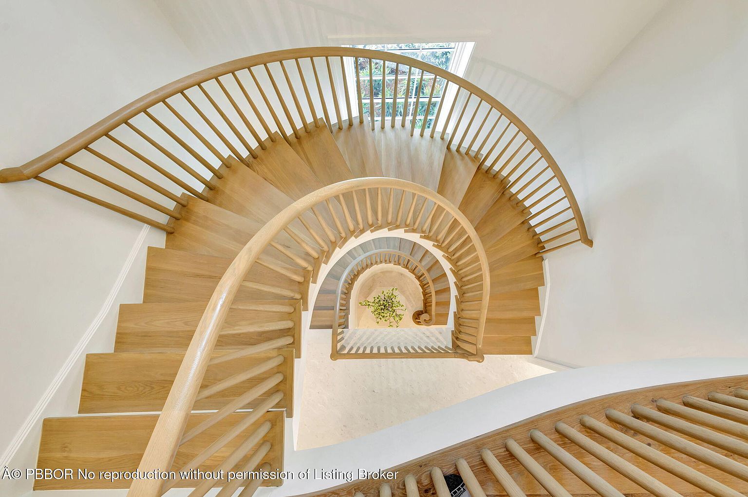 This image showcases a stunning spiral staircase from an overhead perspective, featuring light wood steps and a matching railing. The staircase winds down to a bright, neutral-toned landing with a small plant, creating a visually appealing and elegant architectural detail. Natural light streams in from a window above, enhancing the airy and open feel of the space.