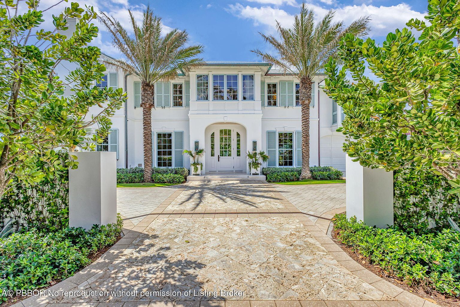 This is a front view of a luxurious two-story white house with symmetrical design elements. The entrance is flanked by two tall palm trees and features a stone pathway leading to the front door. Light blue shutters adorn the windows, and manicured greenery adds to the property's curb appeal, creating an inviting and elegant impression.