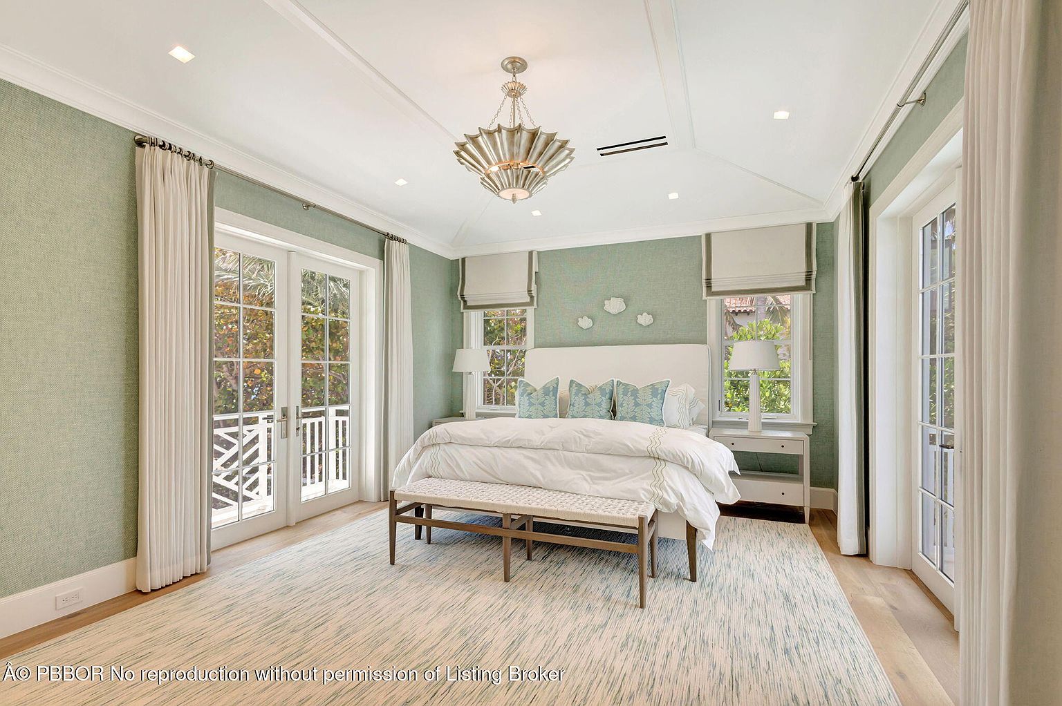 This is a bright and airy primary bedroom featuring a large bed with white linens and blue accent pillows. The room has a soft, textured green wallpaper, white trim, and a decorative chandelier. French doors lead to a balcony, enhancing the room's appeal and natural light.