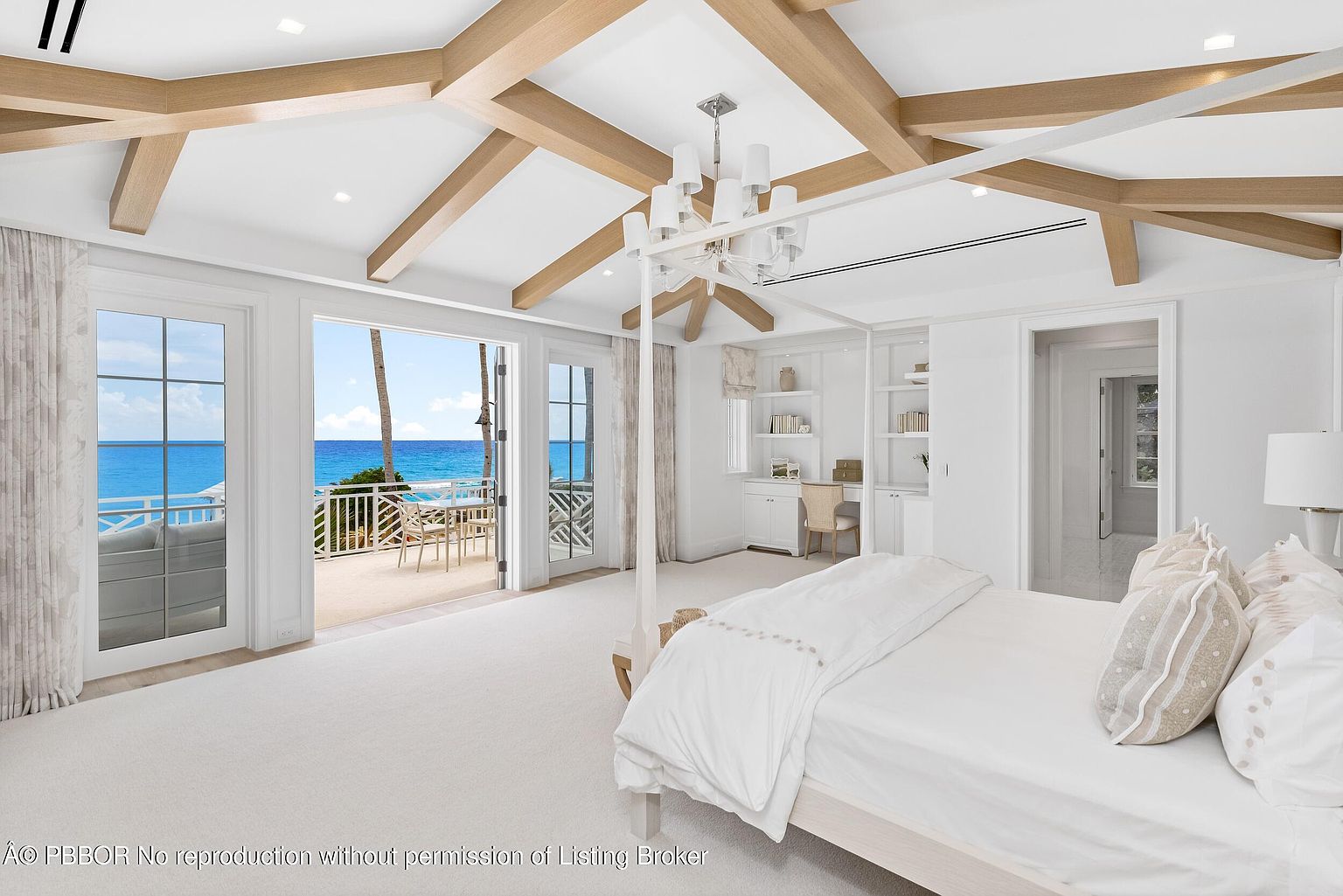 This is a bright and airy primary bedroom with a coastal aesthetic. The room features a four-poster bed, exposed wooden beams on the ceiling, and large doors that open onto a balcony with ocean views. Built-in shelving and a desk area provide functional storage and workspace.