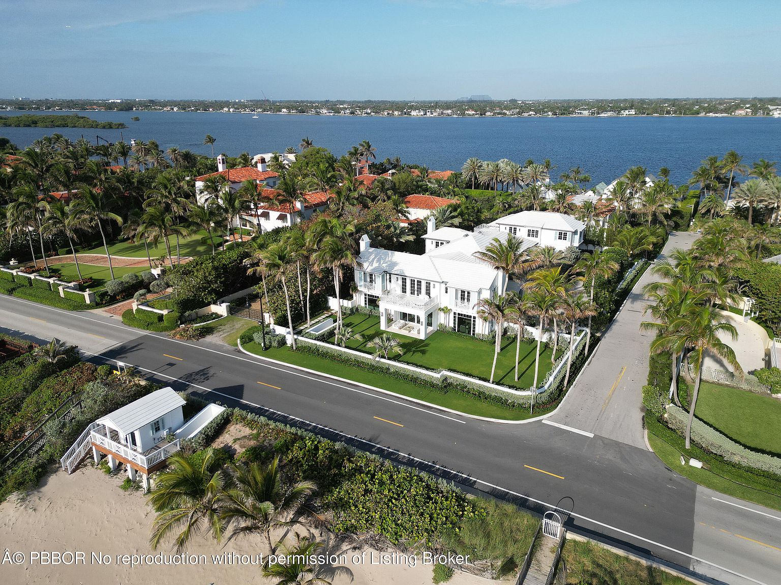 This aerial shot showcases a luxurious waterfront property featuring a large white house with manicured lawns and palm trees. The house is situated on a corner lot with a road on two sides, and the expansive water view in the background adds to the property's appeal. The overall impression is one of high-end coastal living.
