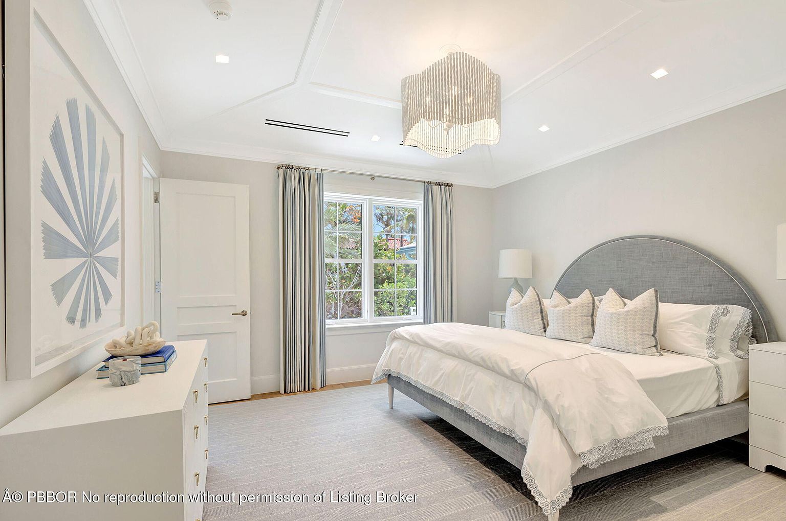 This is a bright and airy primary bedroom featuring a large bed with a gray upholstered headboard and white bedding. The room is decorated in a neutral color palette with blue and gray accents, including striped curtains and a framed art piece. A modern chandelier hangs from the coffered ceiling, and natural light streams in through a window, creating a serene and inviting atmosphere.