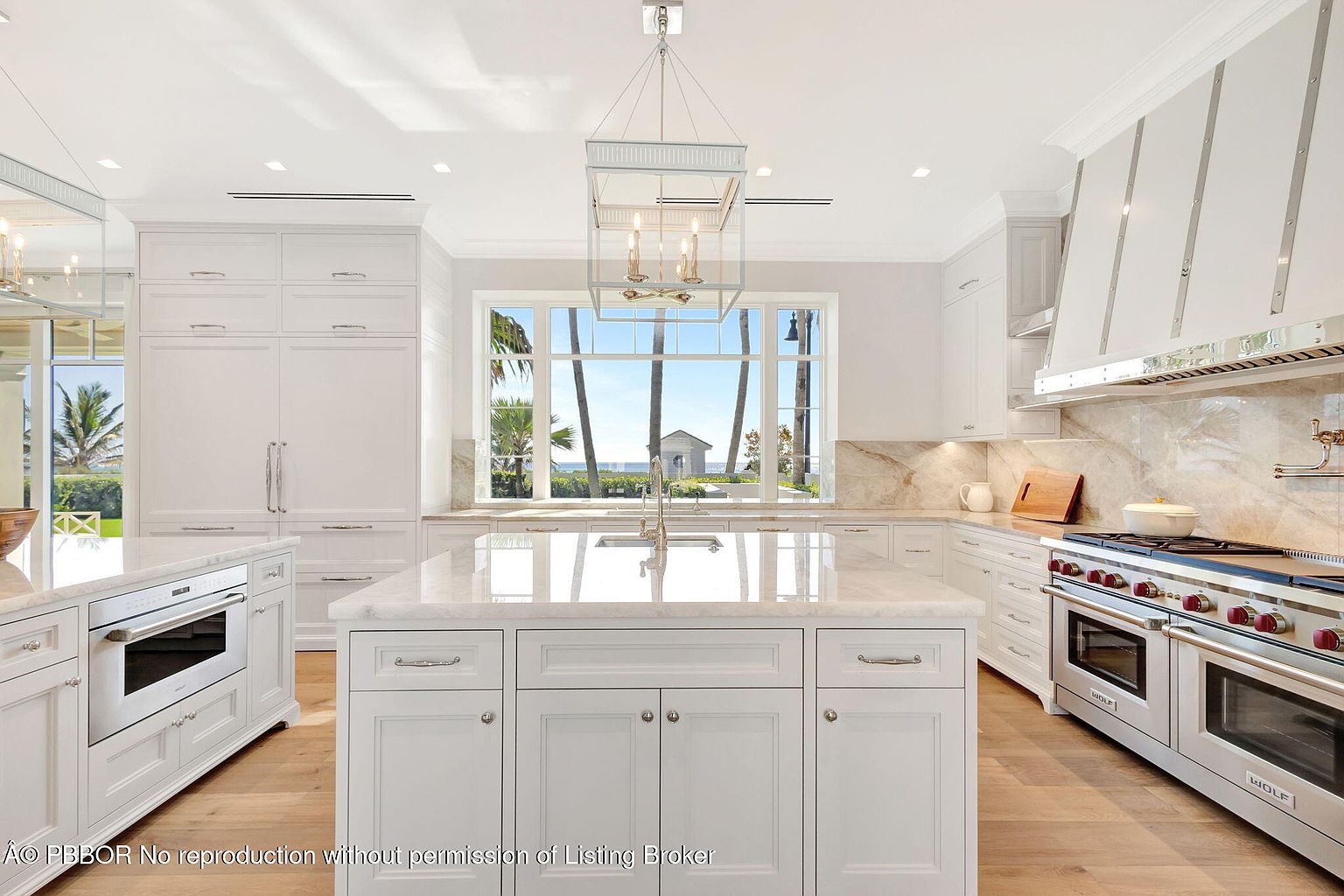 This is a bright and spacious kitchen featuring white cabinetry, a large center island with a marble countertop, and high-end stainless steel appliances including a Wolf range. A large window above the sink offers a view of the ocean and a beach house, enhancing the kitchen's appeal. The overall design is elegant and luxurious, perfect for a high-end real estate listing.